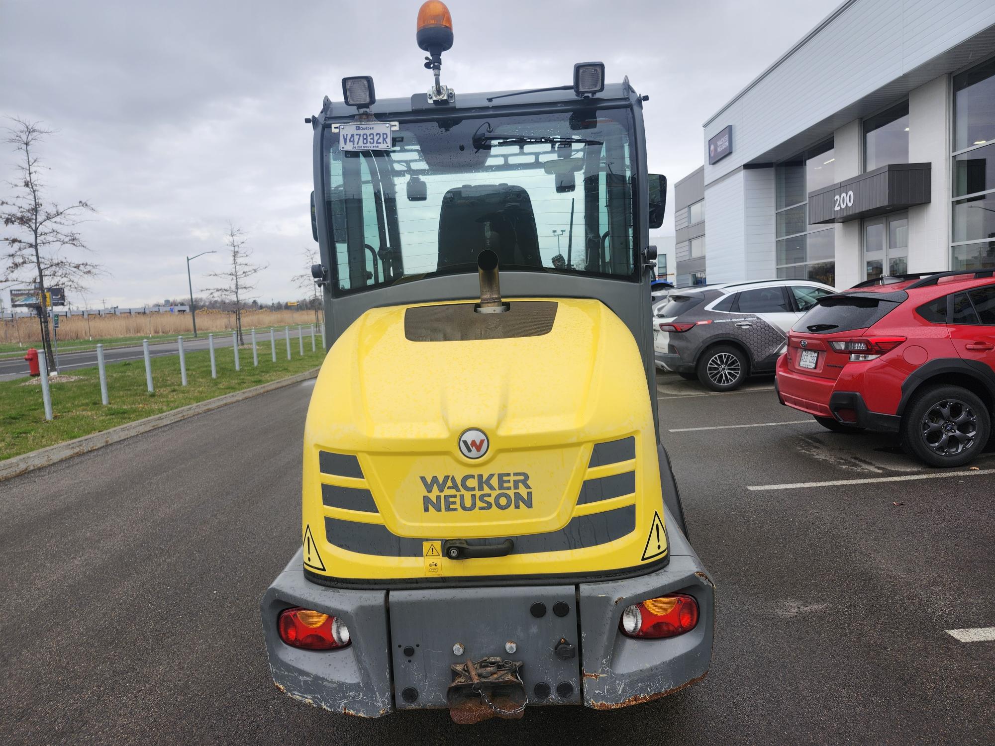 Un véhicule de construction jaune Wacker Neuson est garé sur un terrain pavé à proximité de plusieurs voitures et d'un bâtiment commercial moderne par une journée nuageuse.