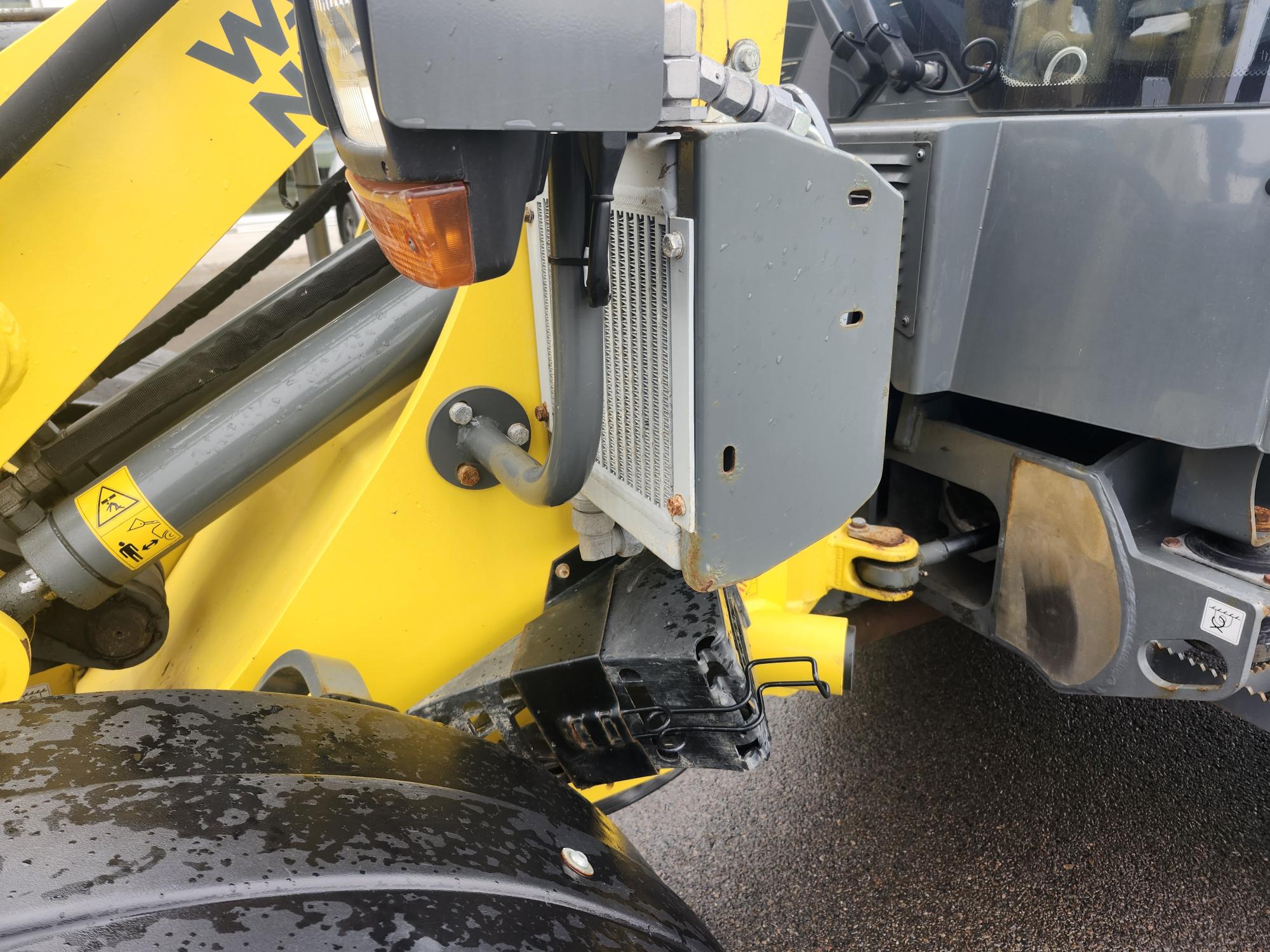 Gros plan d'un engin de chantier jaune, montrant une partie de son bras hydraulique, ses composants métalliques, son radiateur et sa roue avant, avec quelques gouttes d'eau visibles sur les parties noires. Le sol semble mouillé.