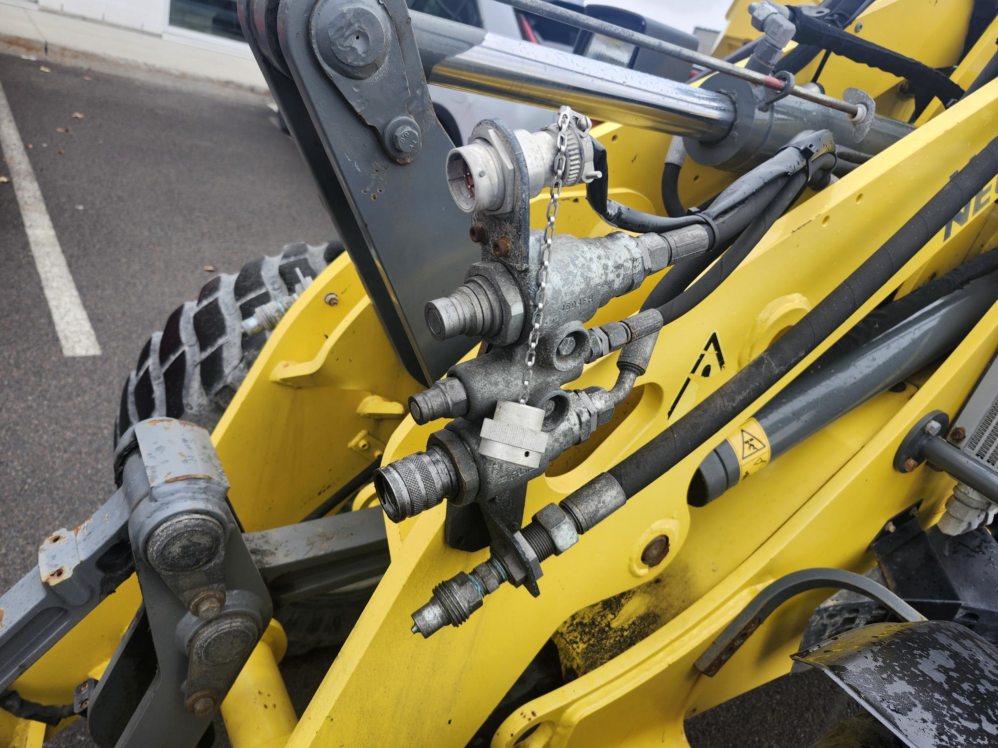 Gros plan sur les raccords et flexibles hydrauliques d'un engin de chantier jaune, avec pièces métalliques, connecteurs et machines visibles. L'engin est stationné sur une surface asphaltée.