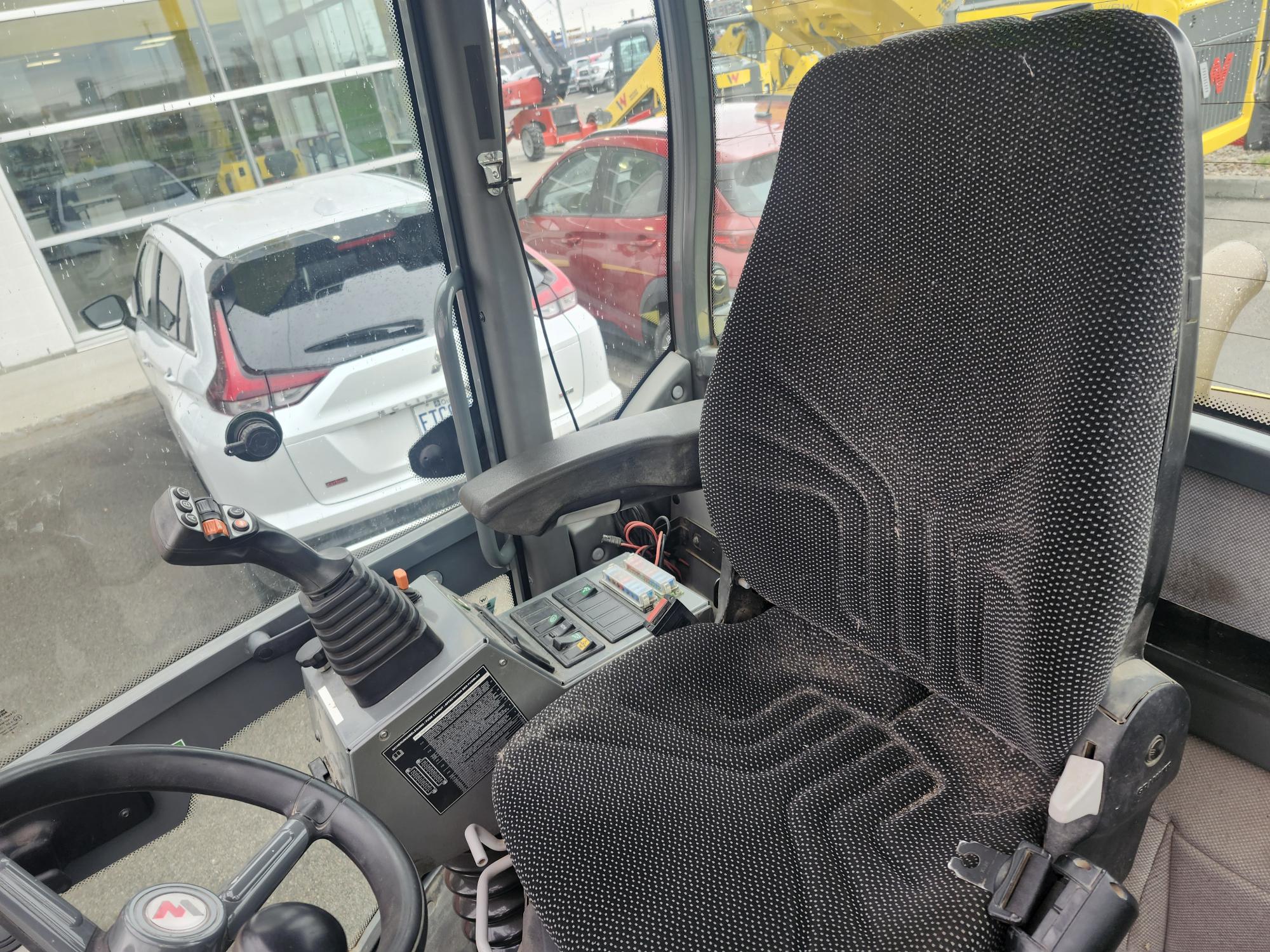 L'image montre le siège conducteur et le panneau de commande d'une cabine d'engin lourd, avec un joystick, des boutons et un volant. Par la fenêtre, on aperçoit des voitures et des engins de chantier stationnés.