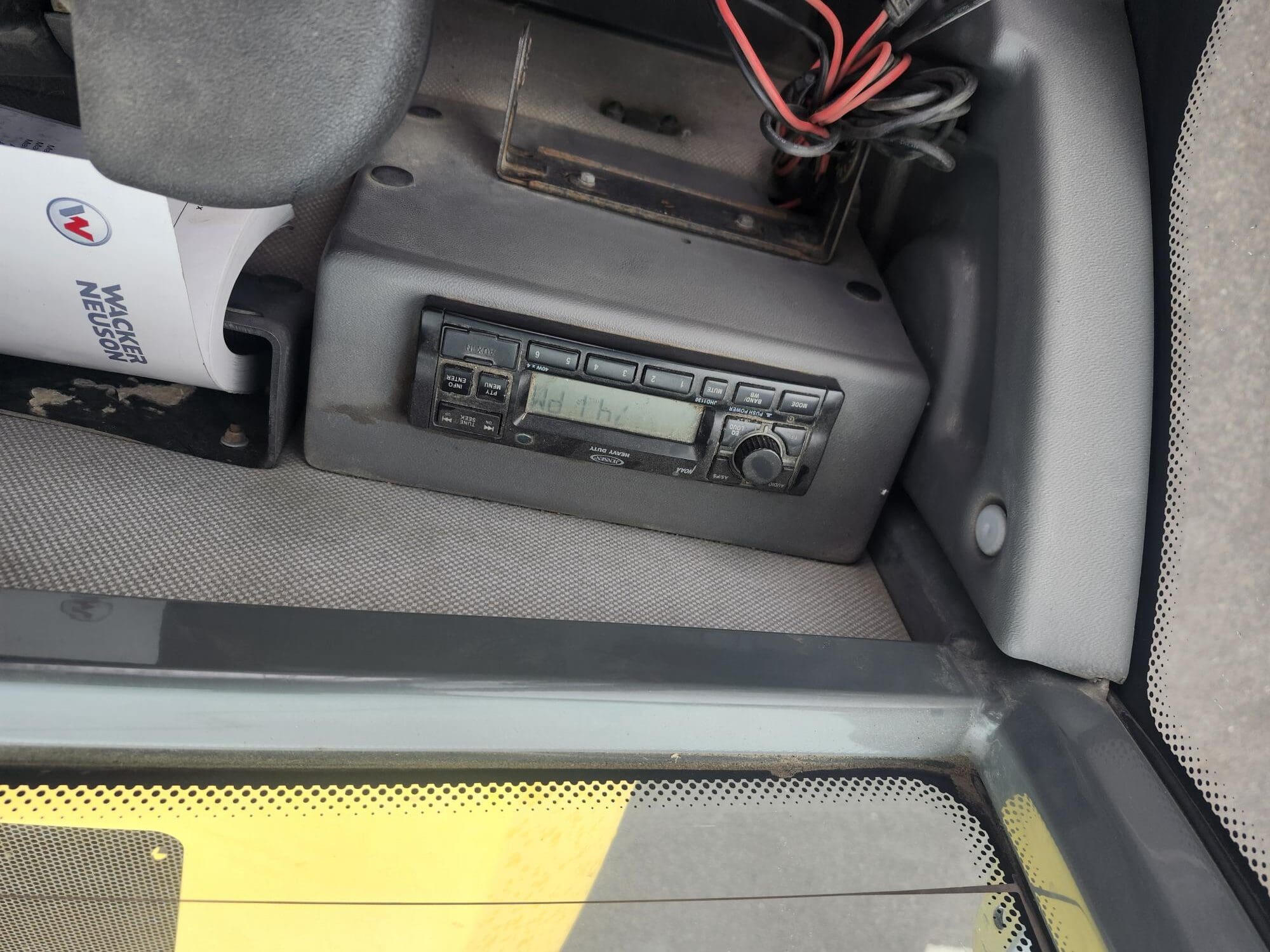 Gros plan d'un autoradio installé sur un tableau de bord, avec des fils apparents au-dessus. À gauche, on peut voir une partie d'un volant et une feuille pliée avec l'inscription « INSPECTION DU VÉHICULE ».