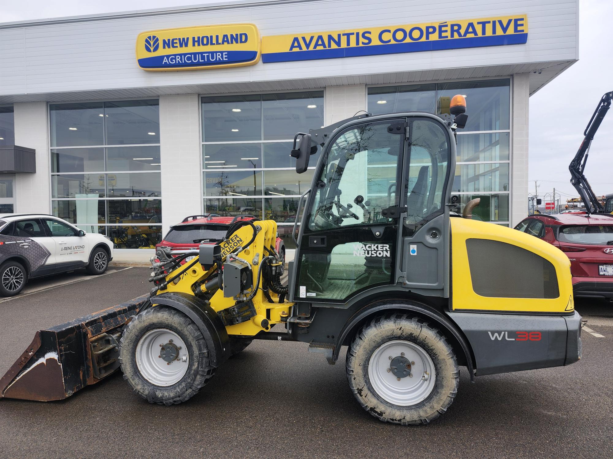 Une chargeuse sur pneus Wacker Neuson WL38 jaune et grise est garée devant un bâtiment de la coopérative agricole New Holland Avantis par temps nuageux. Plusieurs voitures sont garées à proximité.