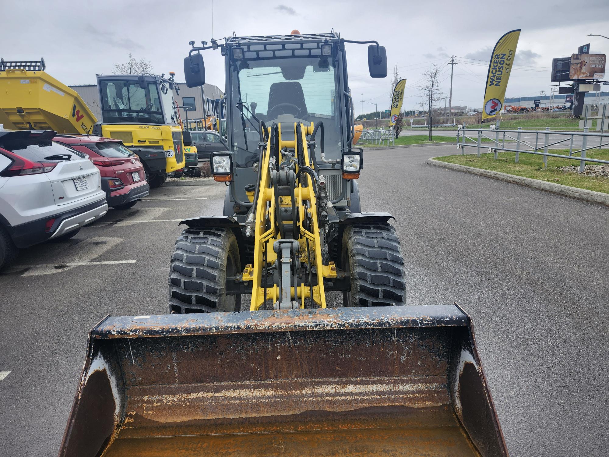 Vue de face d'une chargeuse frontale jaune et noire garée sur le trottoir, godet relevé. Des voitures et autres engins de chantier sont visibles en arrière-plan par temps couvert.
