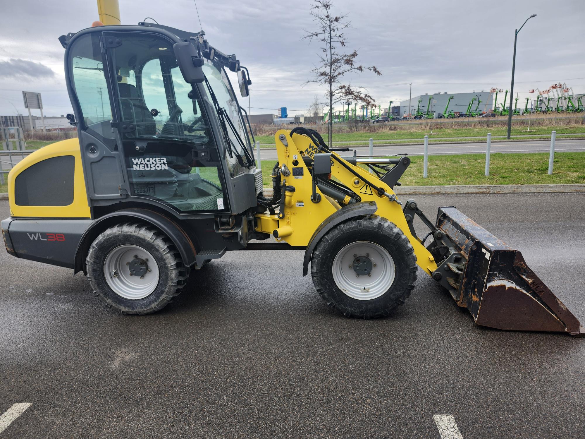 Une chargeuse compacte sur pneus Wacker Neuson WL38 jaune et noire est garée sur une surface asphaltée humide près d'une route, avec des bâtiments industriels et des machines visibles en arrière-plan.