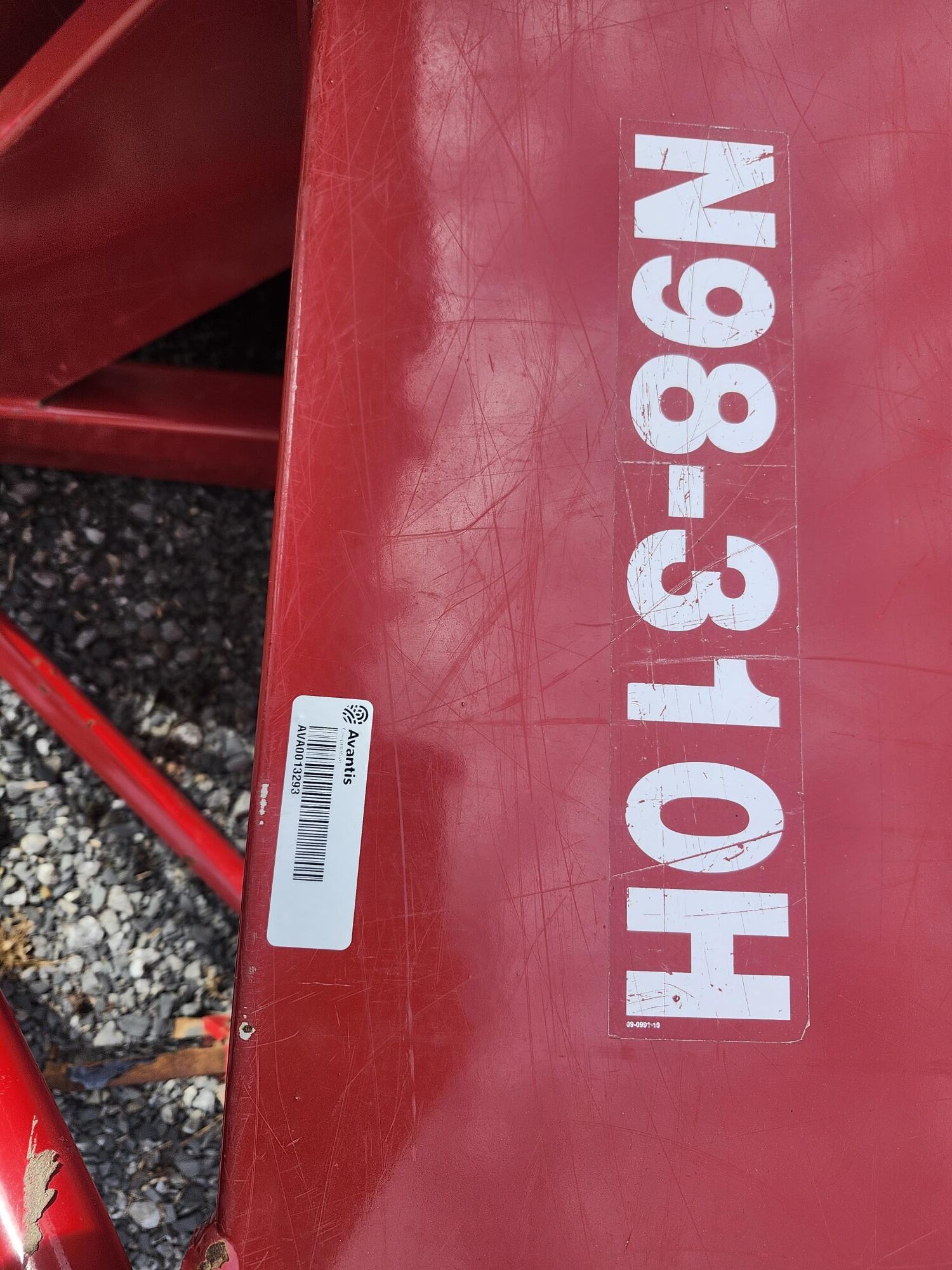 Surface métallique rouge avec inscription blanche au pochoir « N98-310H » et un petit autocollant à code-barres « Amazon » à gauche. Du gravier est visible en arrière-plan.