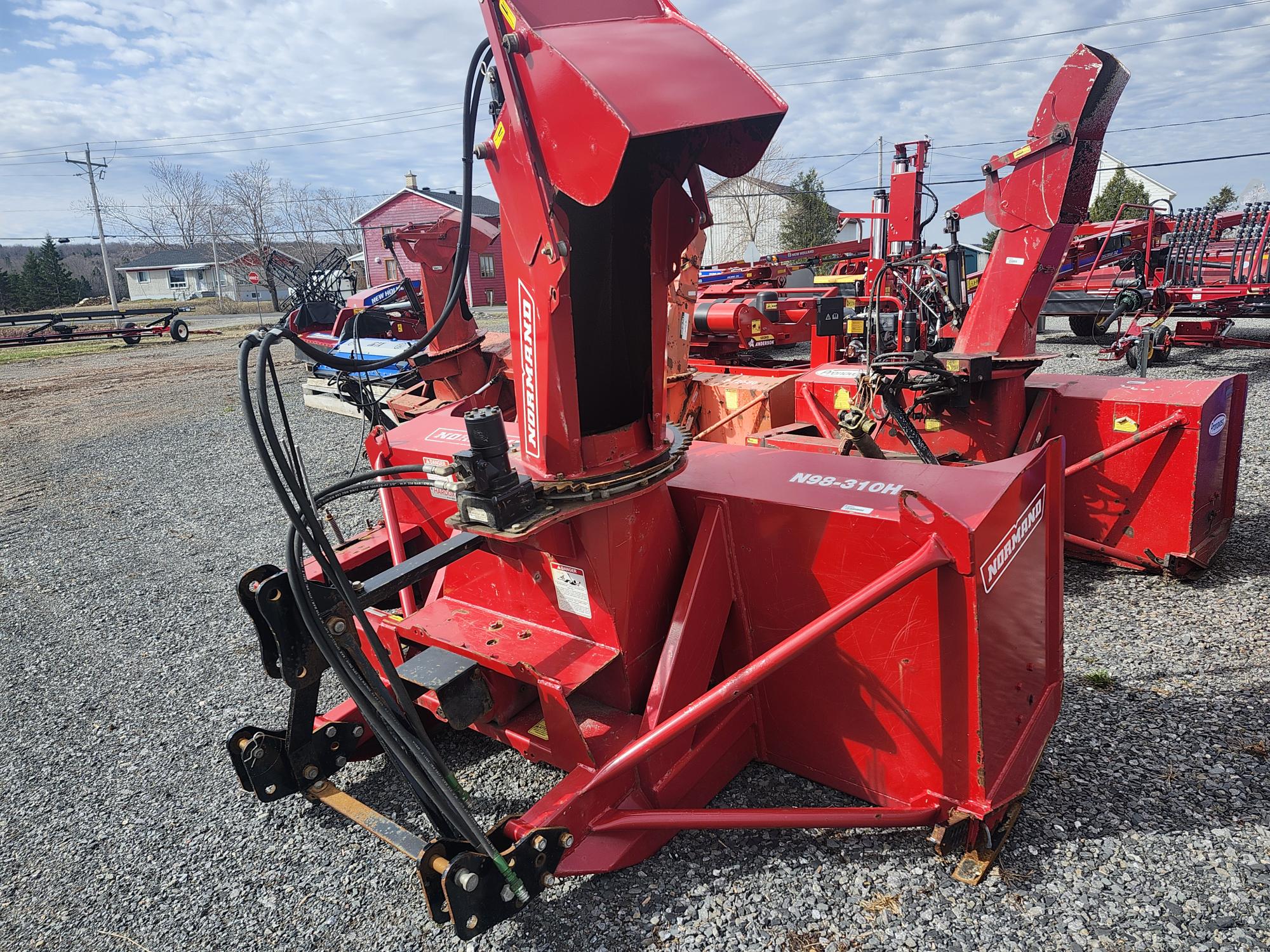 Plusieurs accessoires de souffleuse à neige rouges pour tracteurs sont alignés à l'extérieur sur du gravier. L'équipement comprend des goulottes, des tarières et des composants hydrauliques, avec des bâtiments et des arbres en arrière-plan sous un ciel partiellement nuageux.