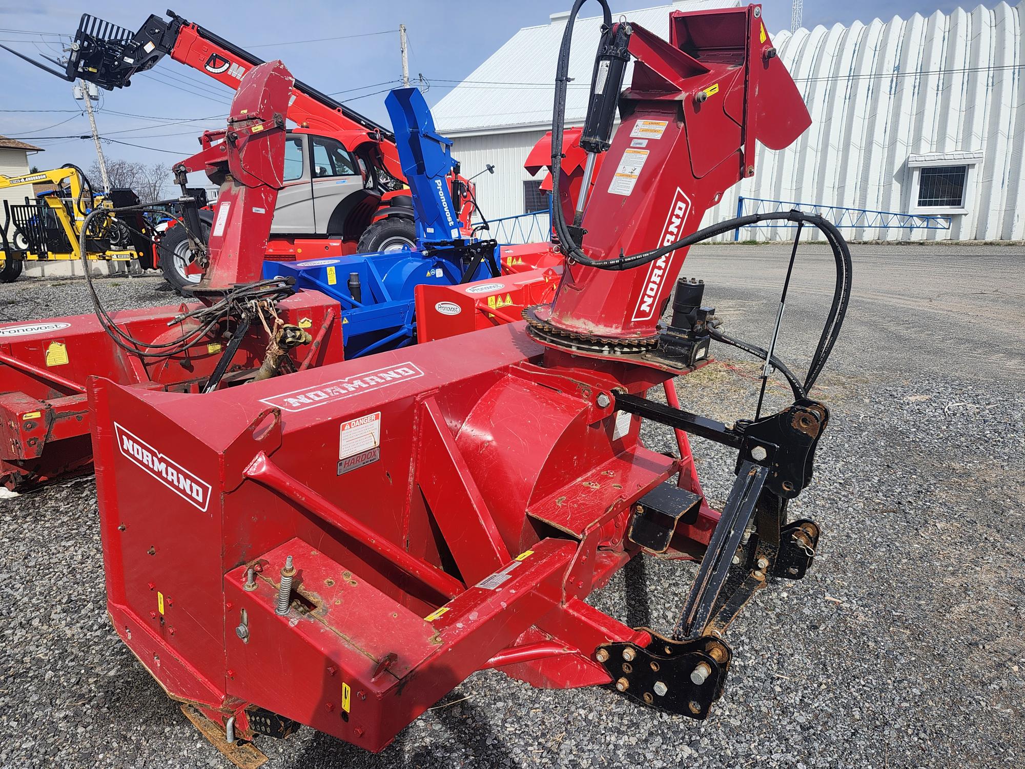Une souffleuse à neige rouge Normand est stationnée sur du gravier, entourée d'autres machines et équipements agricoles, avec un bâtiment en métal et un ciel bleu en arrière-plan.