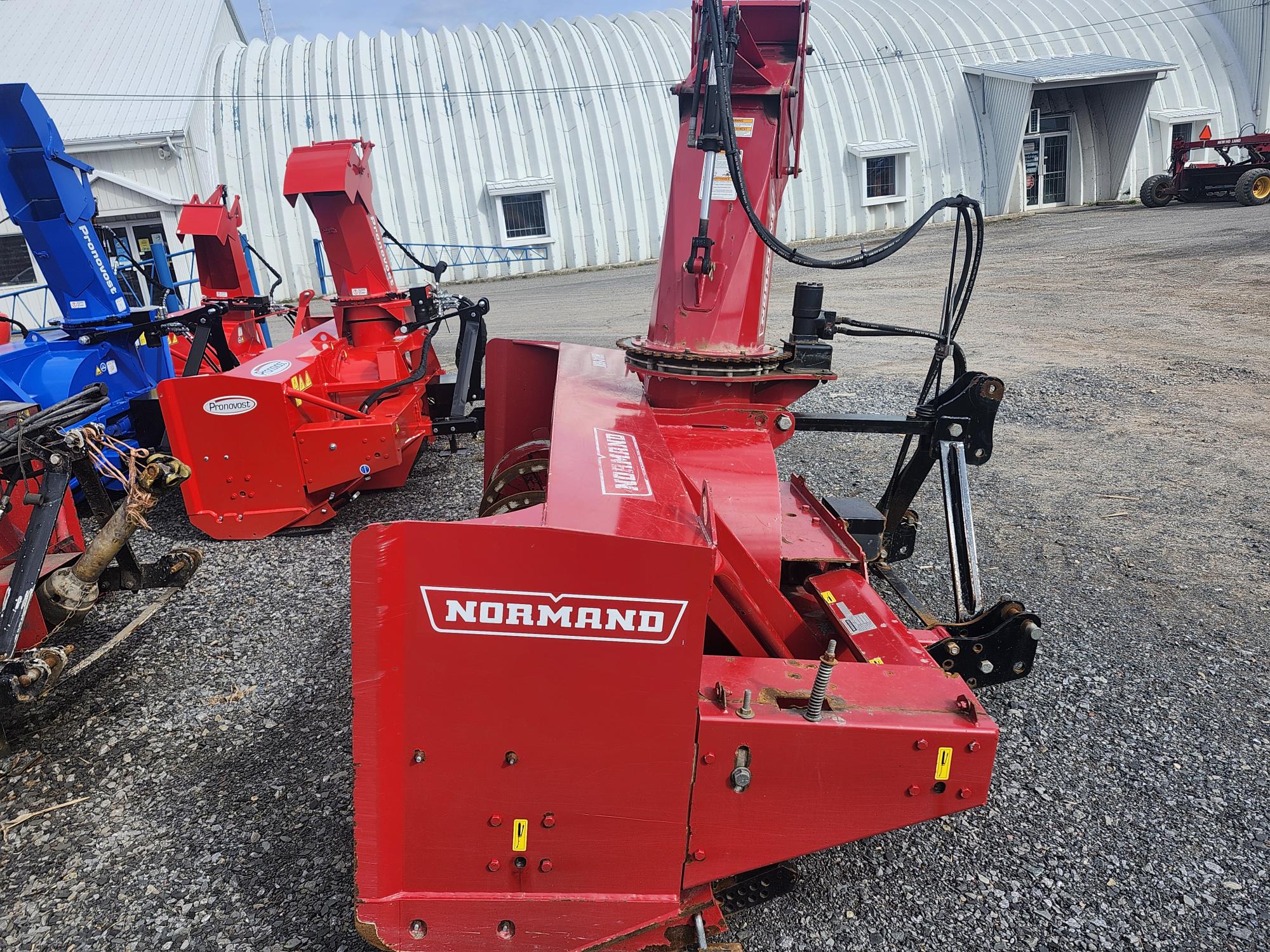 Un gros accessoire de souffleuse à neige rouge avec la marque « Normand » est positionné sur du gravier près d'une machine bleue similaire et d'un bâtiment en tôle ondulée en arrière-plan.