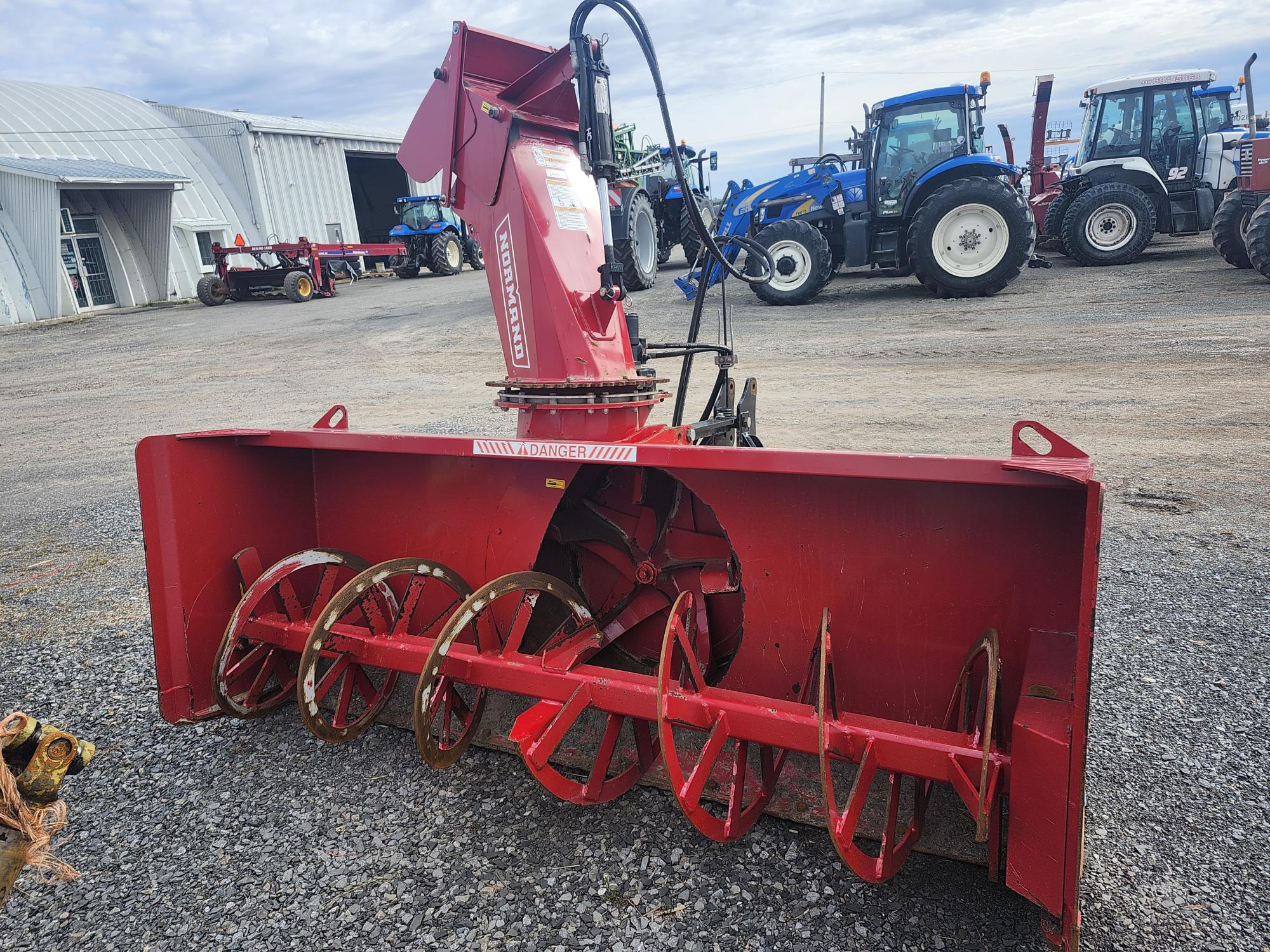 Une grande souffleuse à neige rouge avec tarières est positionnée sur du gravier, avec des tracteurs et d'autres équipements agricoles visibles en arrière-plan près d'une grange en métal.