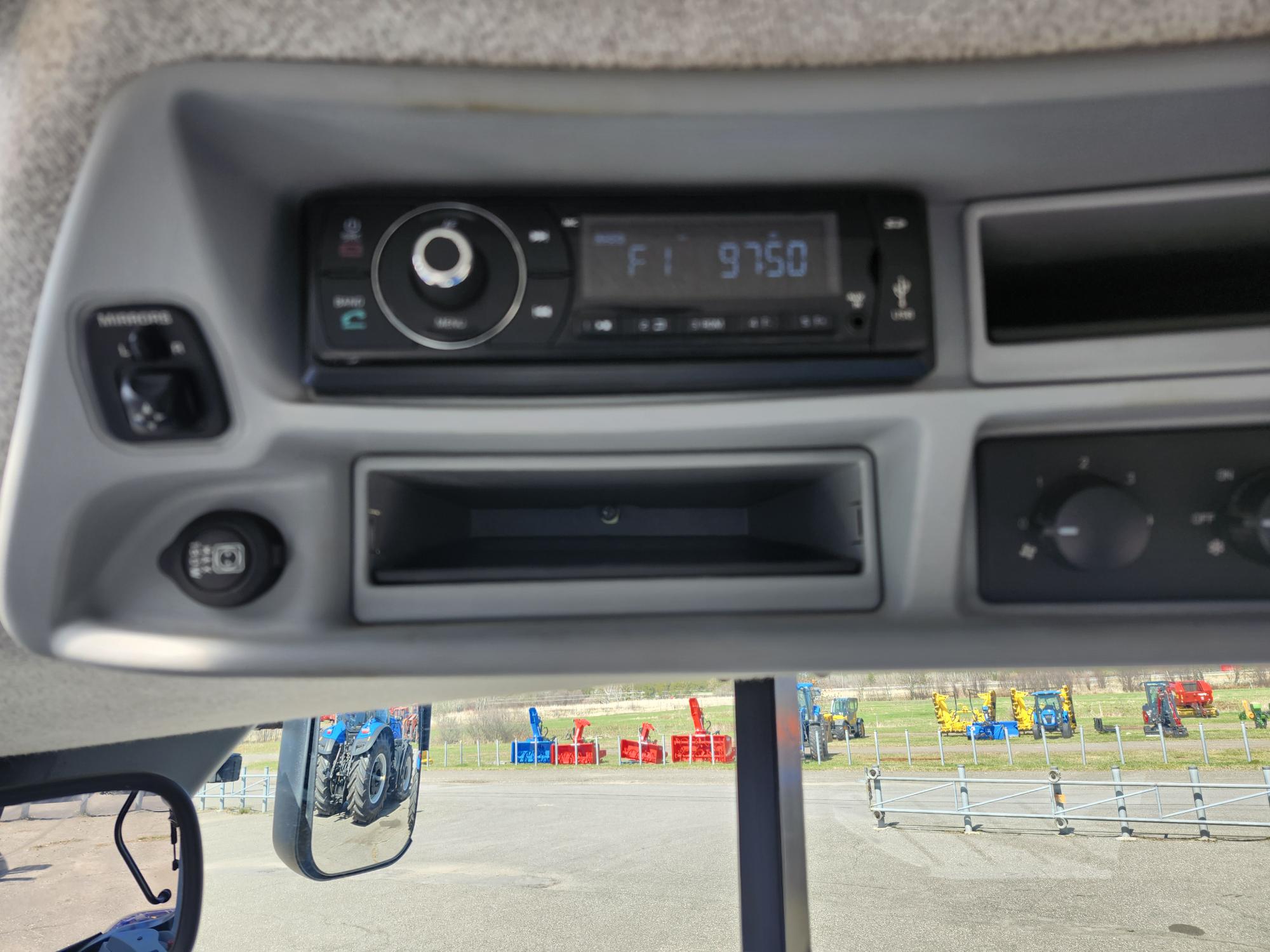Vue rapprochée du tableau de bord d'un véhicule montrant un écran radio numérique réglé sur FM 97,50, des boutons de commande, des interrupteurs et un compartiment de rangement vide avec une vue partielle sur le pare-brise.