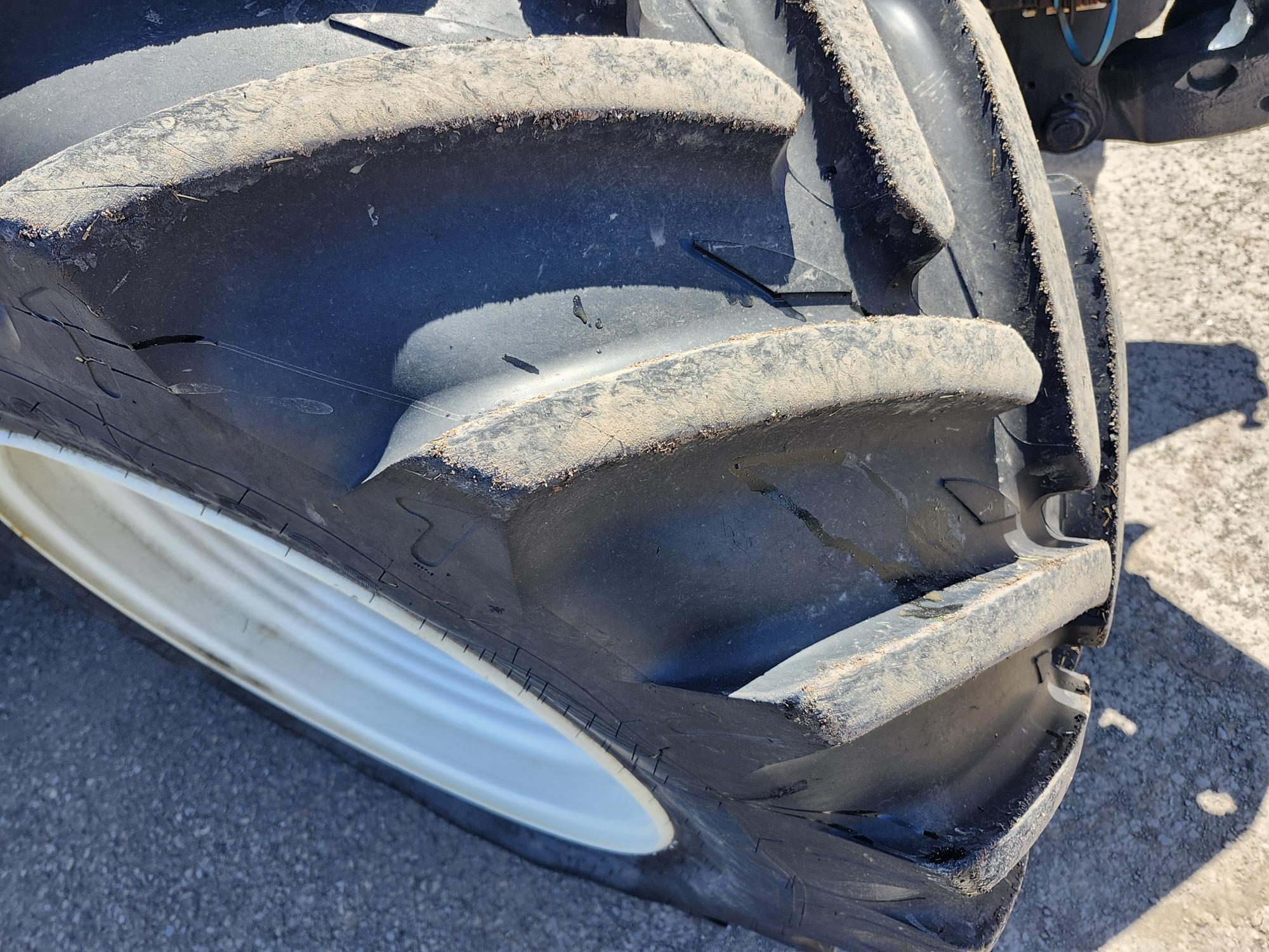 Gros plan d'un gros pneu de tracteur noir avec des bandes de roulement prononcées, montrant de la saleté et de l'usure, positionné sur une surface extérieure rugueuse.