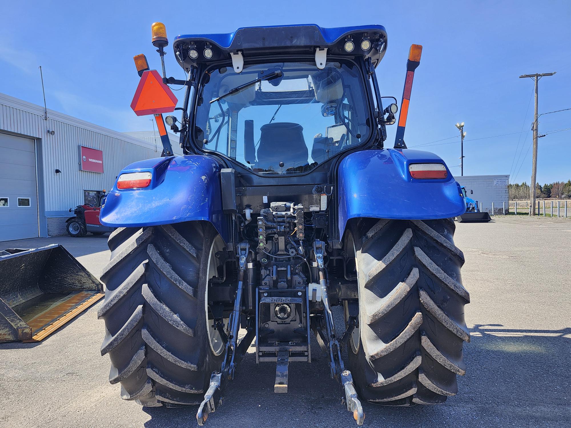 Vue arrière d'un tracteur bleu stationné sur le trottoir, montrant de larges pneus arrière à crampons, des mécanismes d'attelage et un triangle rouge réfléchissant. Un bâtiment et d'autres équipements sont visibles en arrière-plan sous un ciel dégagé.