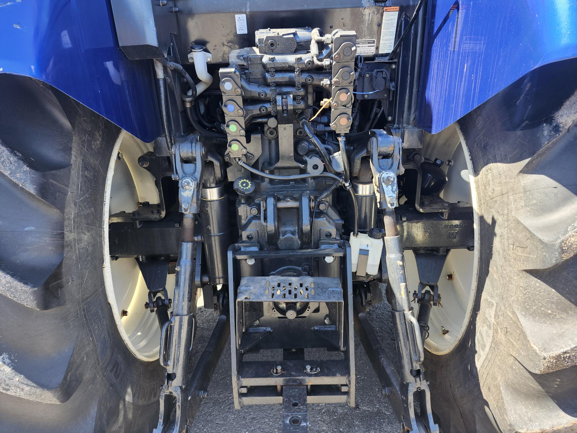 Vue rapprochée du système hydraulique arrière et du mécanisme d'attelage d'un tracteur bleu, montrant divers tuyaux, connecteurs, leviers et composants métalliques entre de gros pneus arrière.