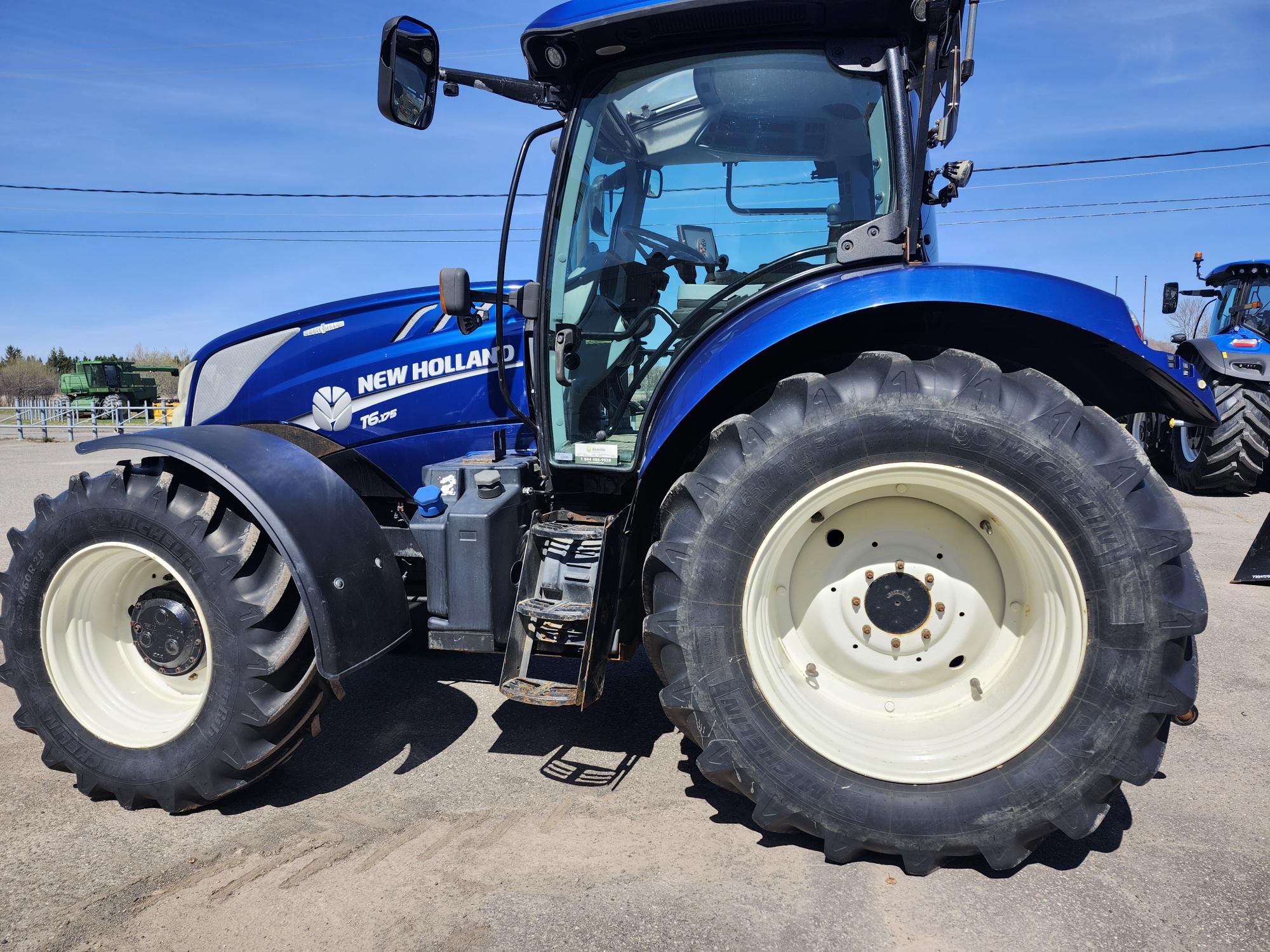 Un tracteur New Holland bleu, équipé de gros pneus noirs, est garé sur une surface pavée sous un ciel bleu clair. La vue latérale met en valeur les roues avant et arrière, ainsi que la cabine.