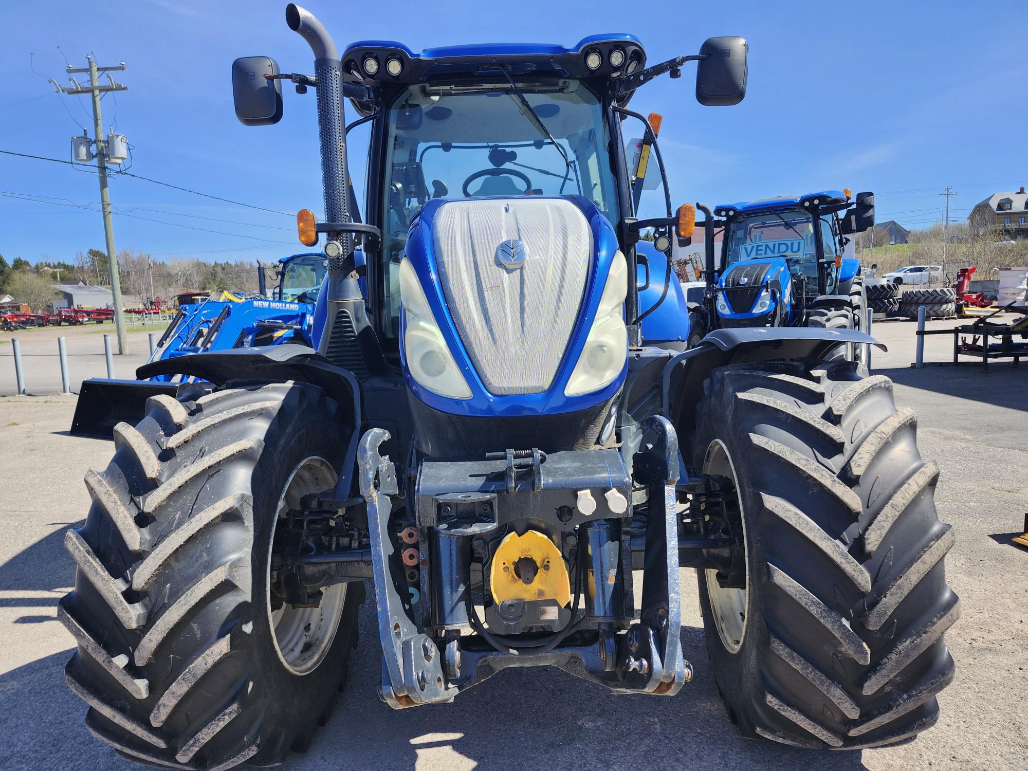 Un gros tracteur bleu avec des pneus larges et robustes est stationné sur un terrain pavé par une journée ensoleillée, avec d'autres équipements agricoles et un panneau « VENDU » (vendu) visibles en arrière-plan.