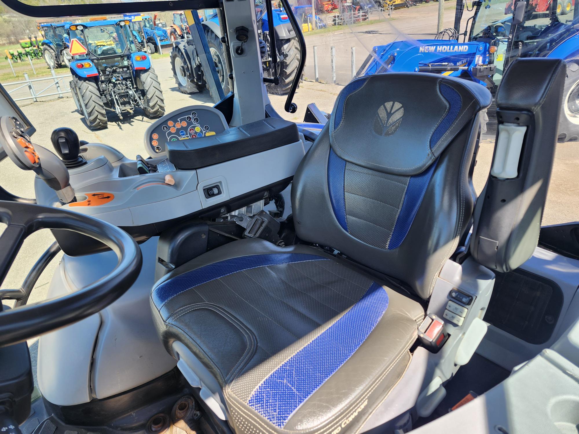 Vue de l'intérieur d'une cabine de tracteur moderne montrant un siège conducteur noir et bleu, un volant, un panneau de commande et une fenêtre, avec d'autres tracteurs bleus visibles à l'extérieur en arrière-plan.
