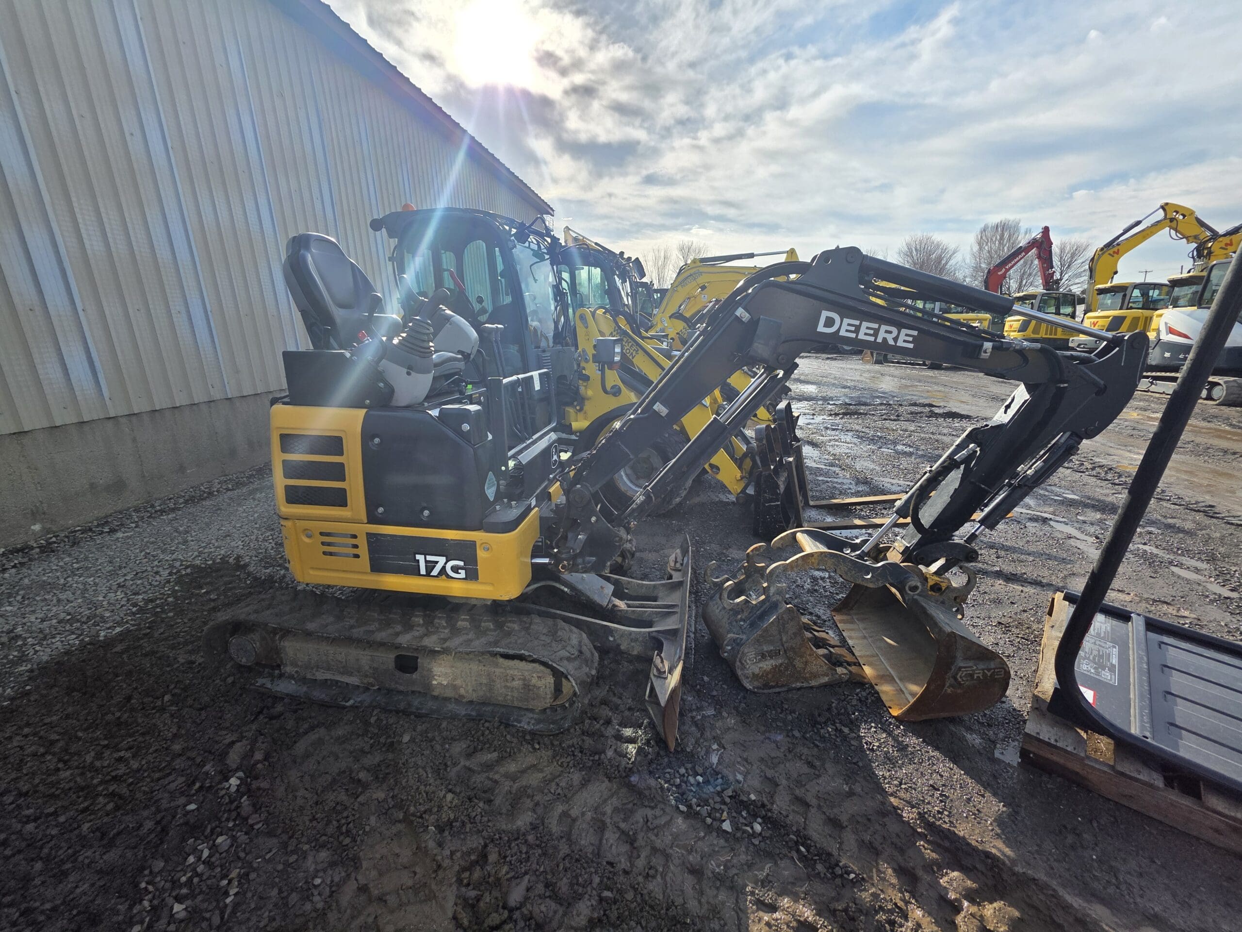 Une mini-pelle jaune portant l'inscription « Deere 17G » est garée sur un terrain de construction boueux, à côté d'autres engins. Le soleil brille intensément à travers un ciel nuageux, projetant des ombres sur le bâtiment et les équipements à proximité.