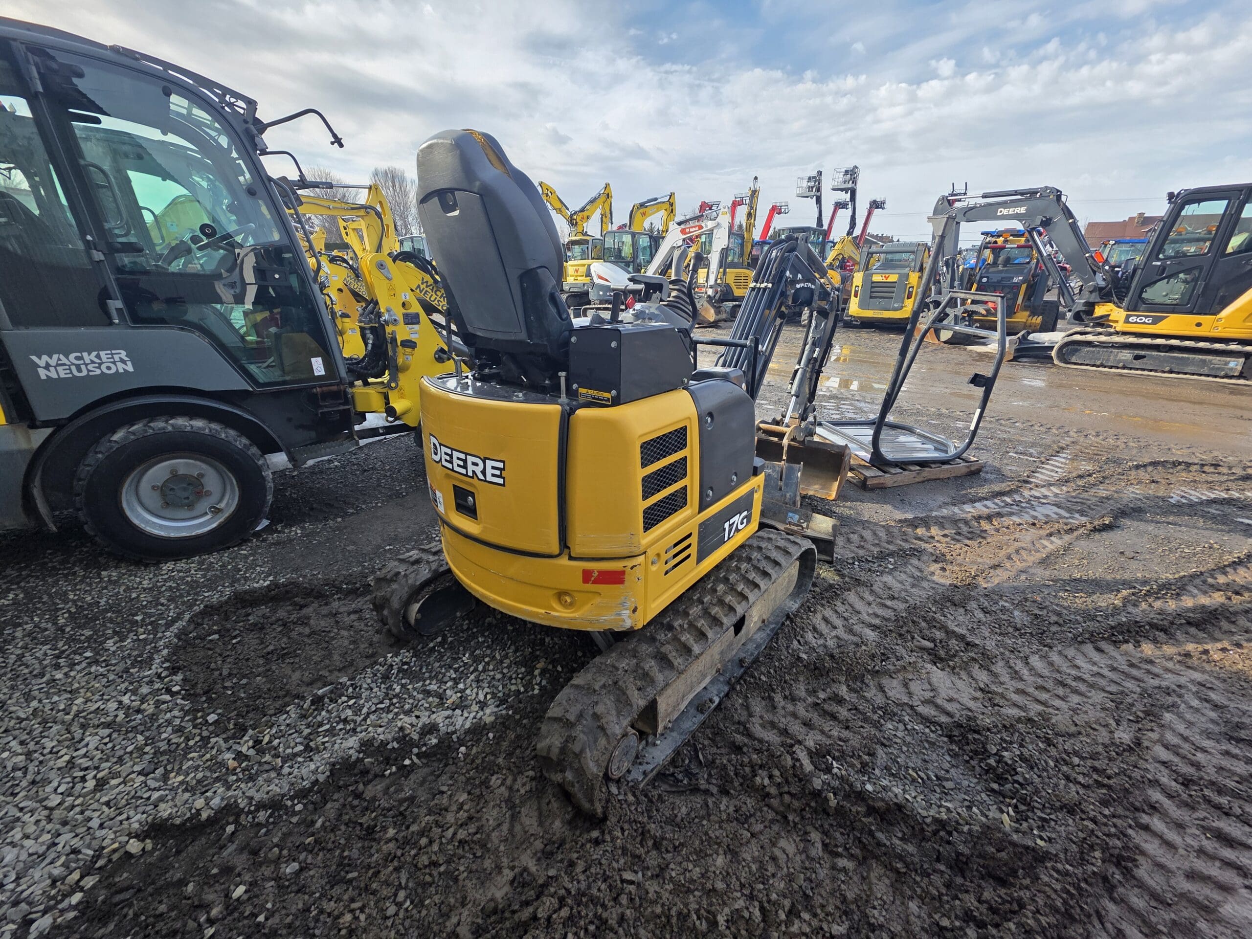 Une mini-pelle jaune portant le logo « DEERE » est garée sur un terrain en gravier boueux. Plusieurs autres engins de chantier sont visibles en arrière-plan. La scène se déroule sous un ciel partiellement nuageux.