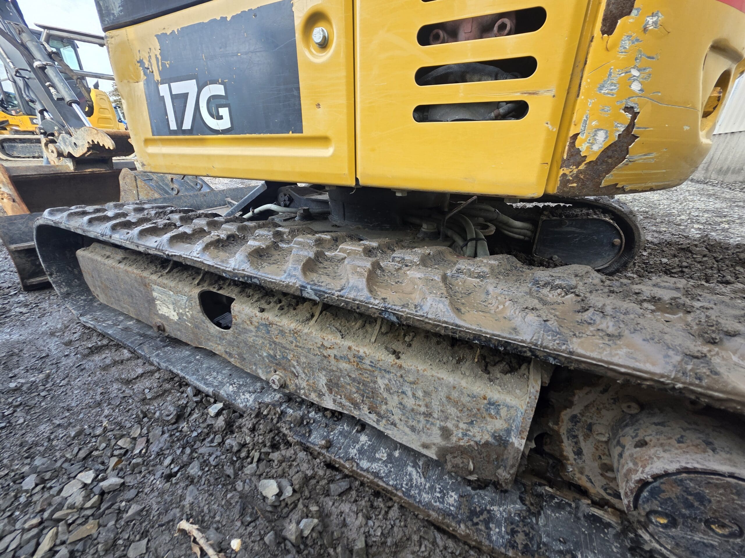 Gros plan sur la section de chenilles jaune et boueuse d'un engin de chantier, étiquetée « 17G ». Les chenilles sont poussiéreuses et l'engin est stationné sur un sol rocailleux. L'attention est portée sur la bande de roulement et les pièces mécaniques visibles en dessous.