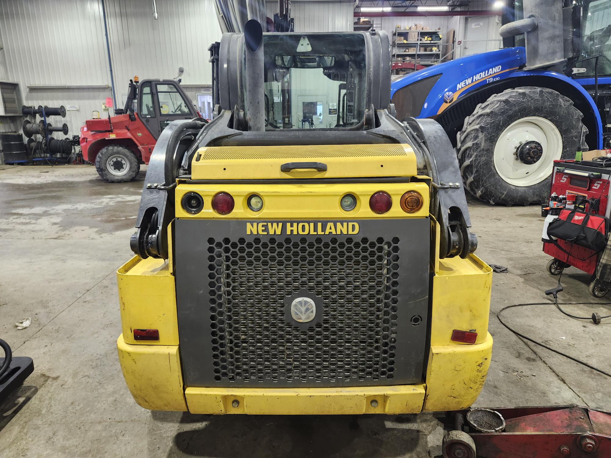 Chargeuse compacte New Holland jaune garée à l'intérieur, dos à la route. Chariot élévateur rouge et tracteur bleu en arrière-plan. Divers outils et équipements sont disséminés dans le grand atelier.