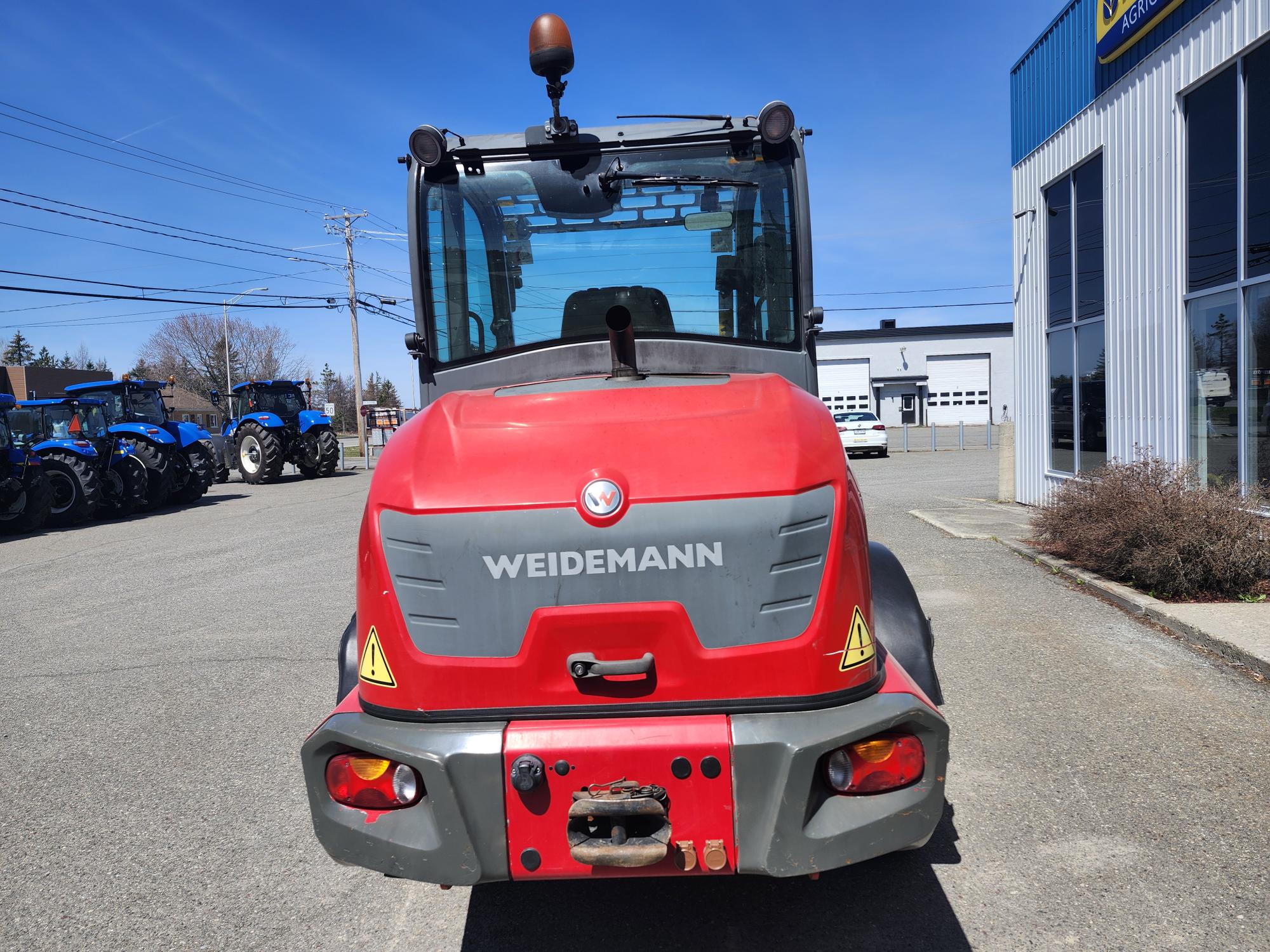 Un véhicule agricole Weidemann rouge est garé à l'extérieur sur un terrain pavé, vu de l'arrière. D'autres tracteurs et un bâtiment métallique sont visibles en arrière-plan sous un ciel bleu clair.
