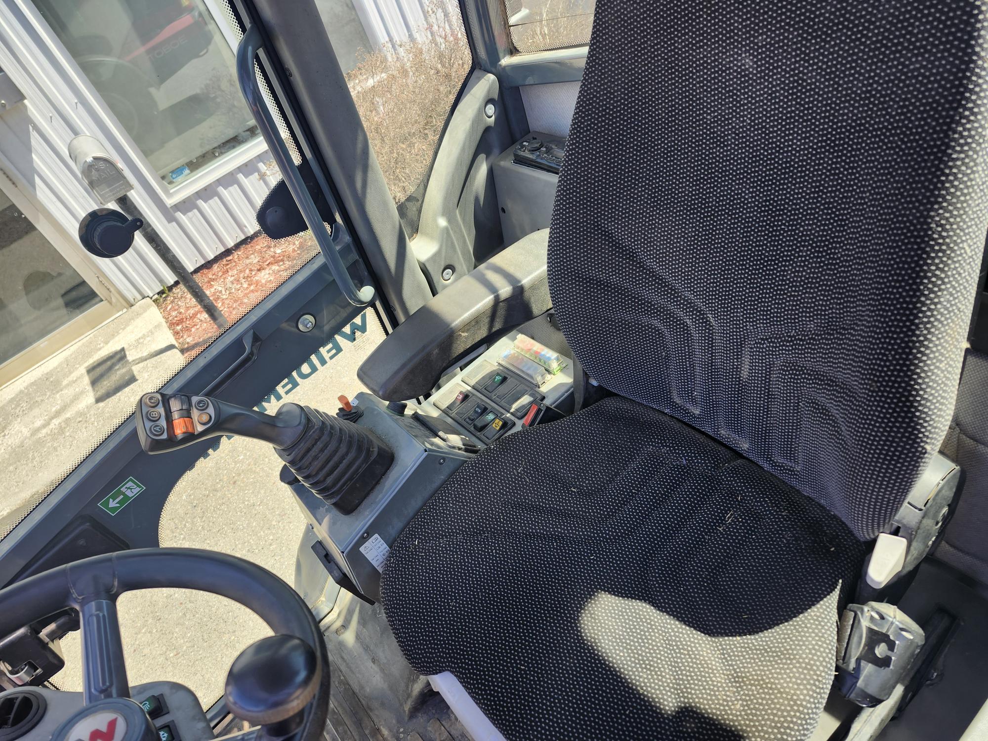 Gros plan de l'intérieur d'un véhicule, montrant le siège conducteur, le volant et le panneau de commande avec ses multiples boutons et son joystick. Le soleil éclaire le siège et les commandes.
