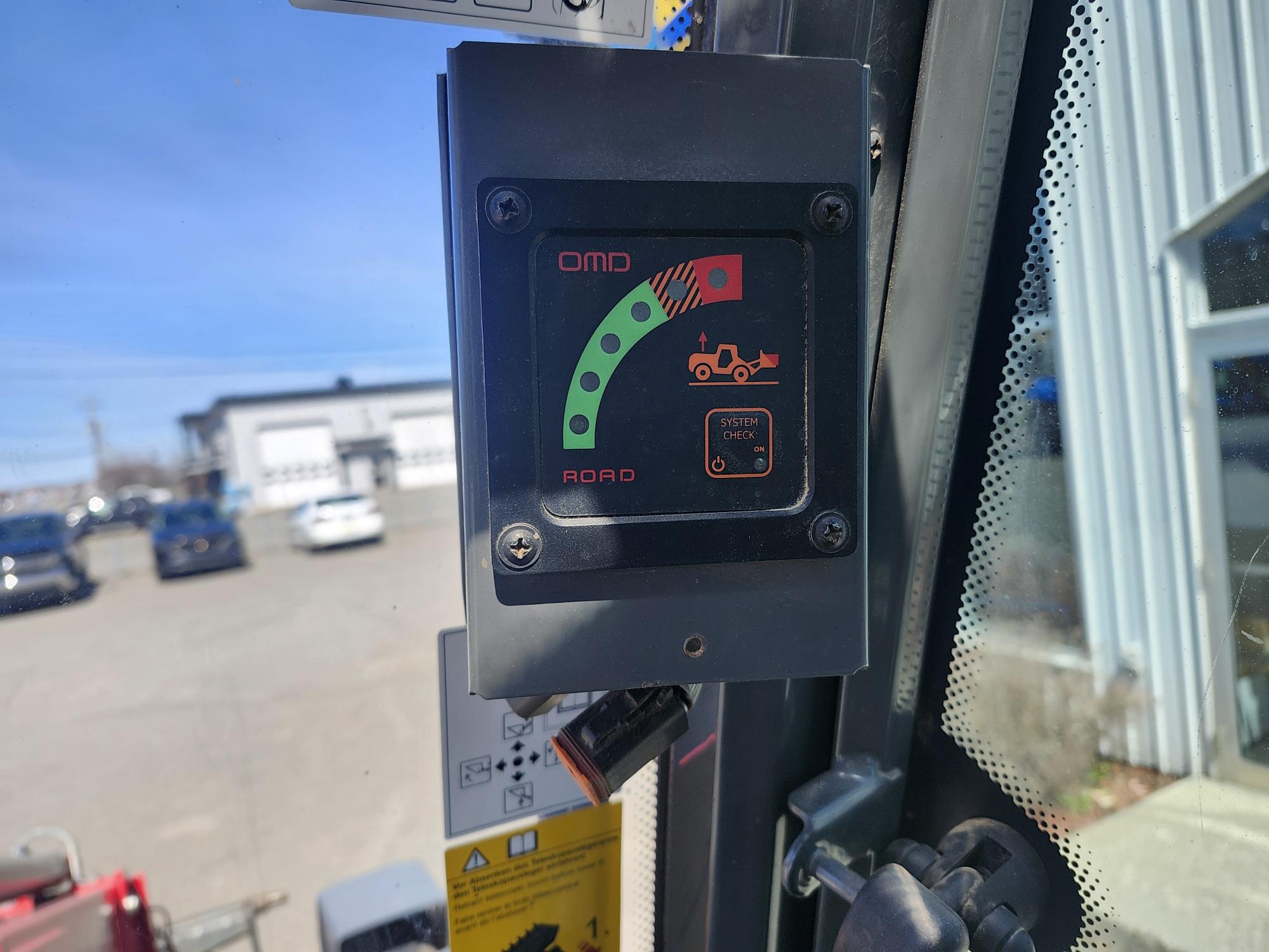 Gros plan d'un écran de tableau de bord de véhicule montrant un indicateur d'avertissement avec un graphique d'un camion et un point d'exclamation rouge, étiqueté « VÉRIFICATION DU SYSTÈME », à l'intérieur d'une cabine avec des bâtiments et des voitures garées à l'extérieur.