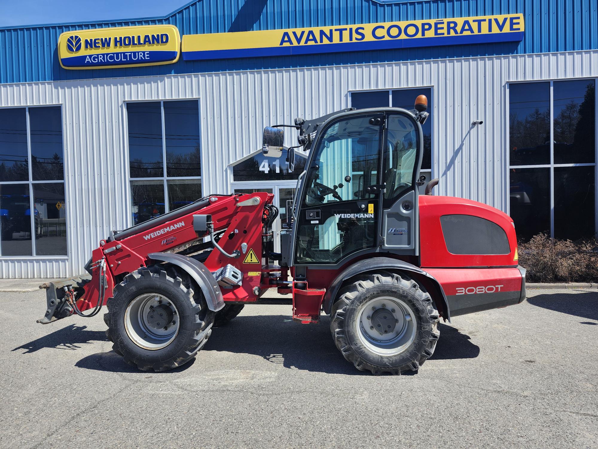 Une chargeuse rouge Weidemann 3080T est garée à l'extérieur d'un bâtiment bleu et blanc avec des panneaux « New Holland Agriculture » et « Avantis Cooperative » au-dessus de l'entrée.