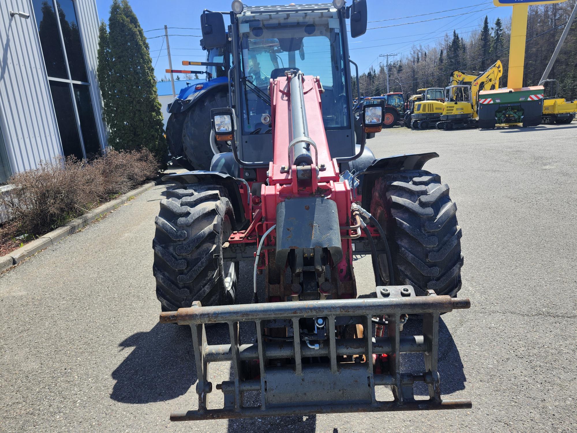 Vue de face d'un chariot télescopique rouge avec de gros pneus garé sur le trottoir, entouré d'autres véhicules de construction et industriels par une journée ensoleillée.