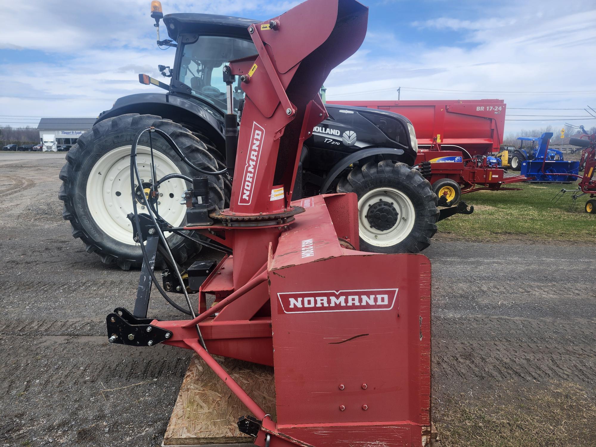 Un accessoire de souffleuse à neige Normand rouge est positionné devant un tracteur New Holland noir sur une surface de gravier, avec d'autres équipements agricoles visibles en arrière-plan sous un ciel partiellement nuageux.