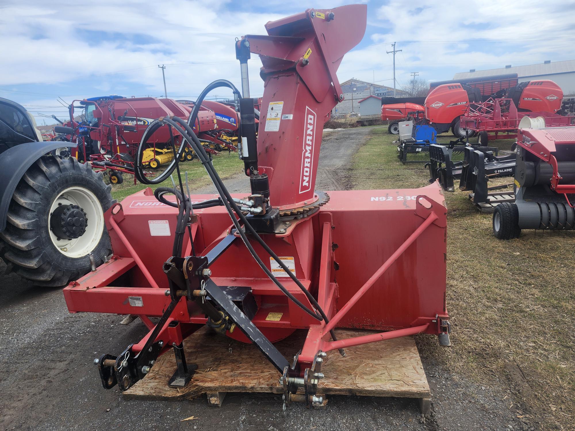 Une souffleuse à neige rouge Normand N92-280 est exposée à l'extérieur sur une palette en bois, entourée d'autres équipements et machines agricoles, avec un ciel nuageux au-dessus.