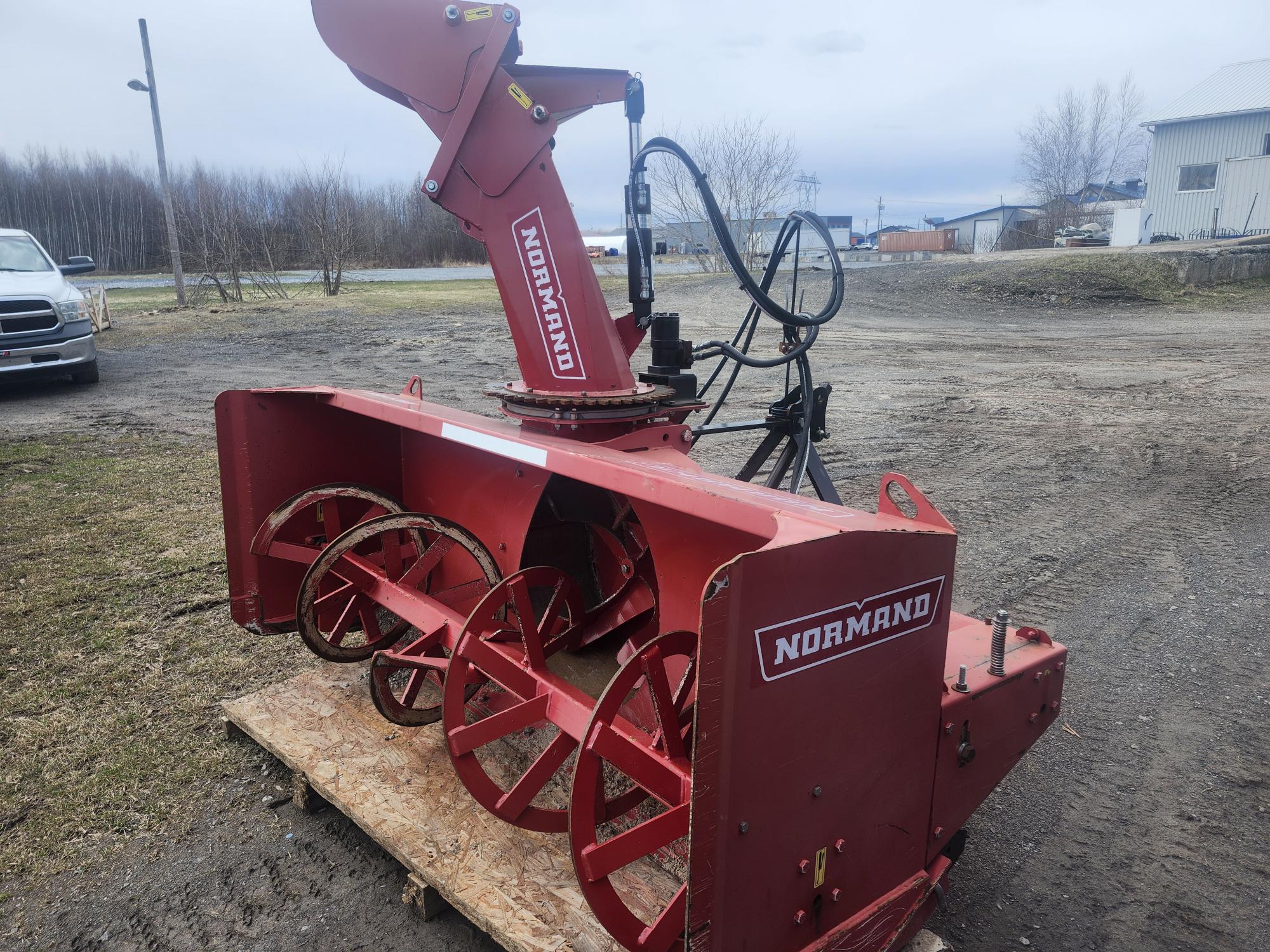Une grande souffleuse à neige Normand rouge est posée à l'extérieur sur une palette, sa goulotte étant relevée et sa tarière exposée. Un camion et des bâtiments industriels sont visibles en arrière-plan sous un ciel nuageux.