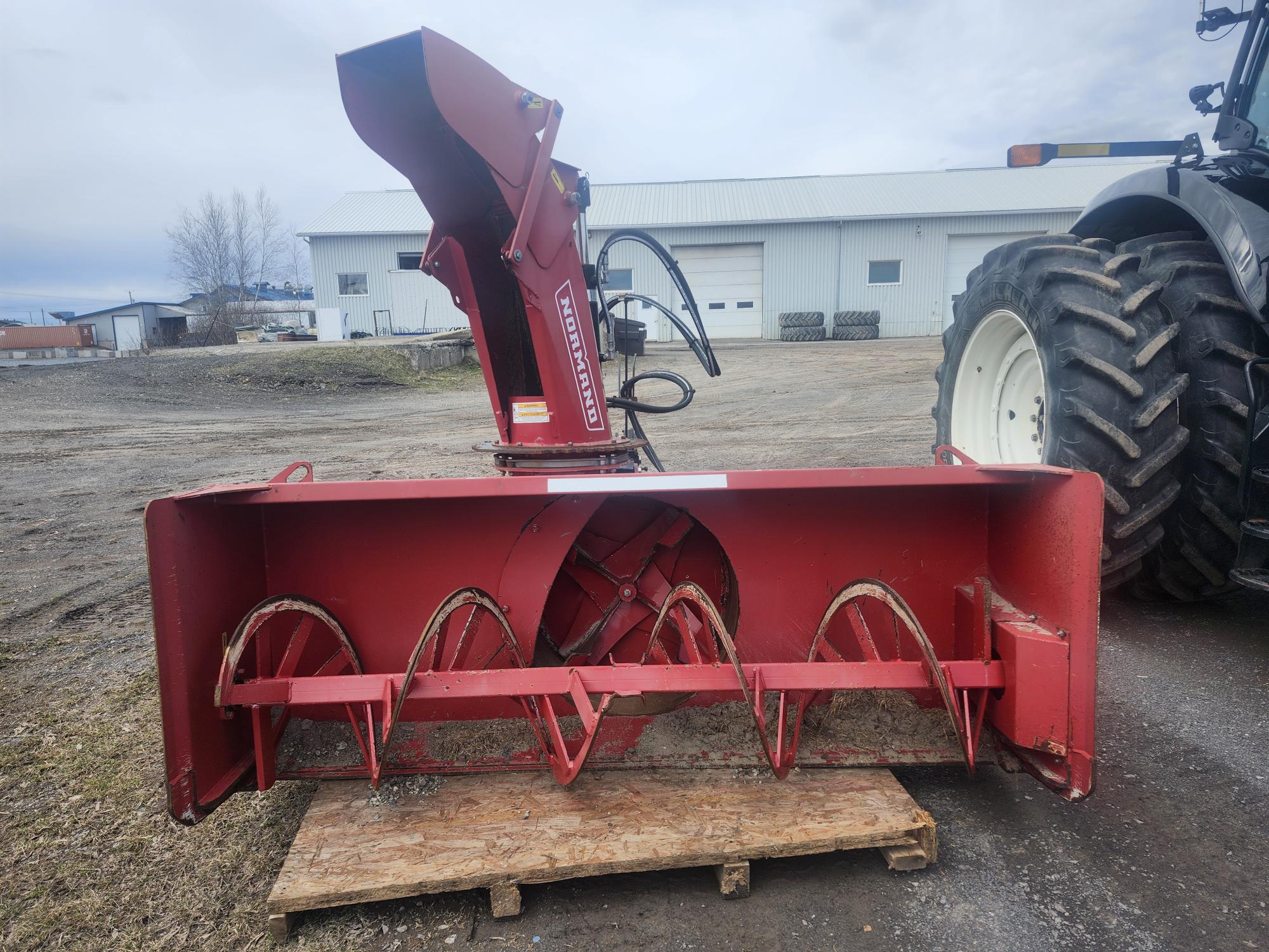 Un accessoire de souffleuse à neige rouge est posé sur une palette en bois à l'extérieur, avec une roue de tracteur partiellement visible à droite et un bâtiment industriel en arrière-plan.