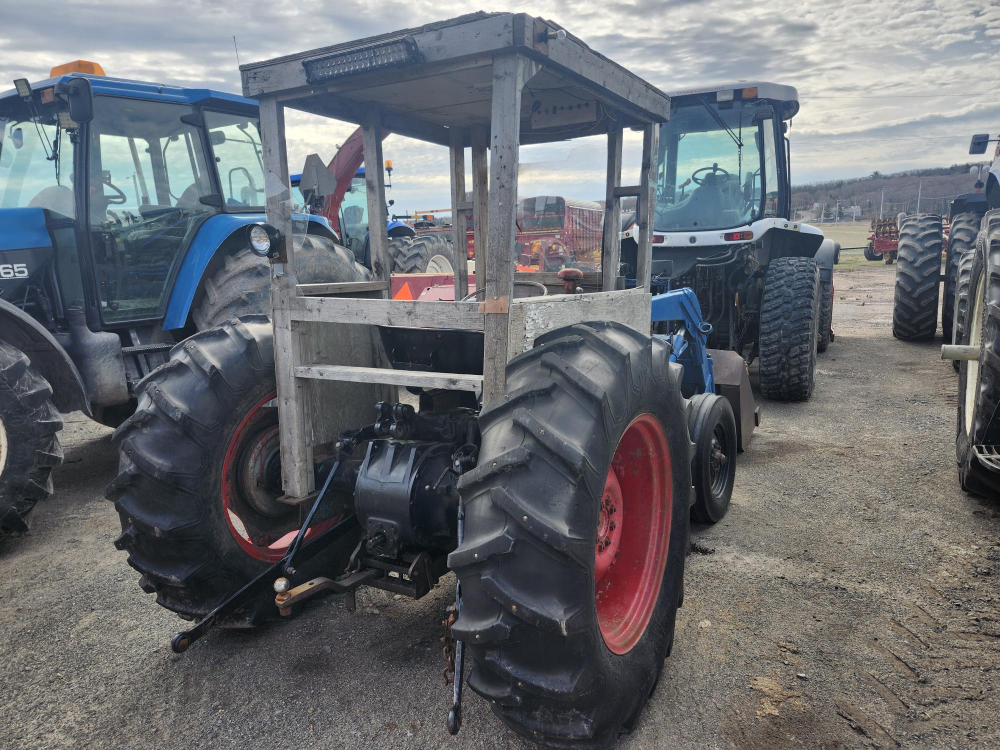 Un tracteur bleu avec de gros pneus arrière et une cabine en bois faite maison est garé parmi d'autres tracteurs agricoles sur un terrain en gravier sous un ciel nuageux.
