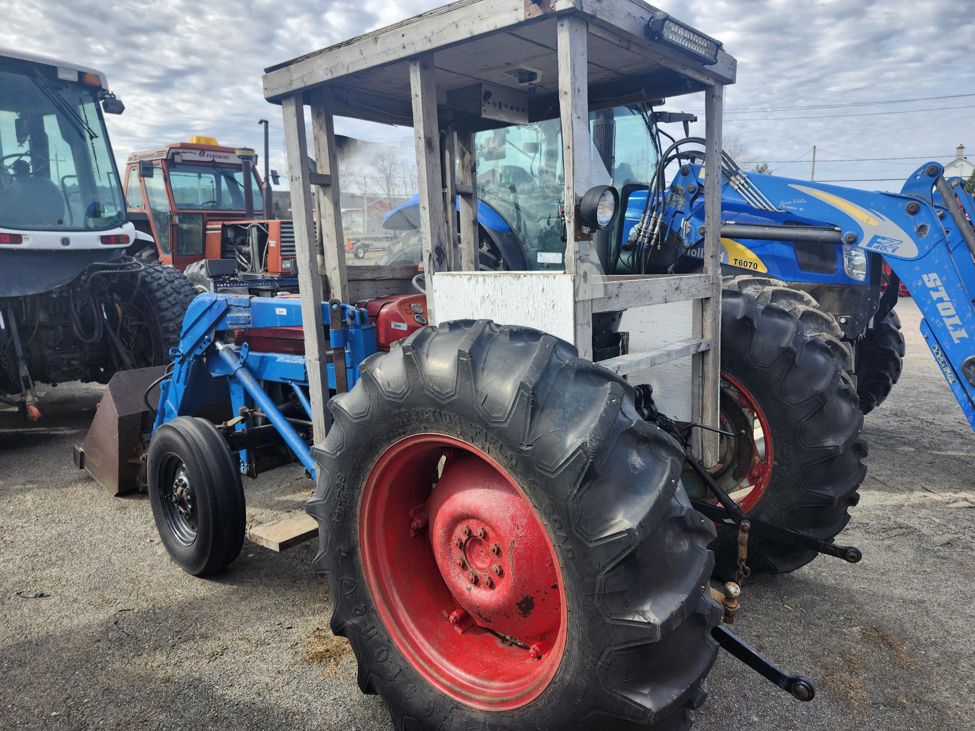 Vue rapprochée d'un vieux tracteur bleu avec de gros pneus noirs et des jantes rouges, doté d'une cabine blanche partiellement fermée, garé à côté d'autres tracteurs sous un ciel nuageux.