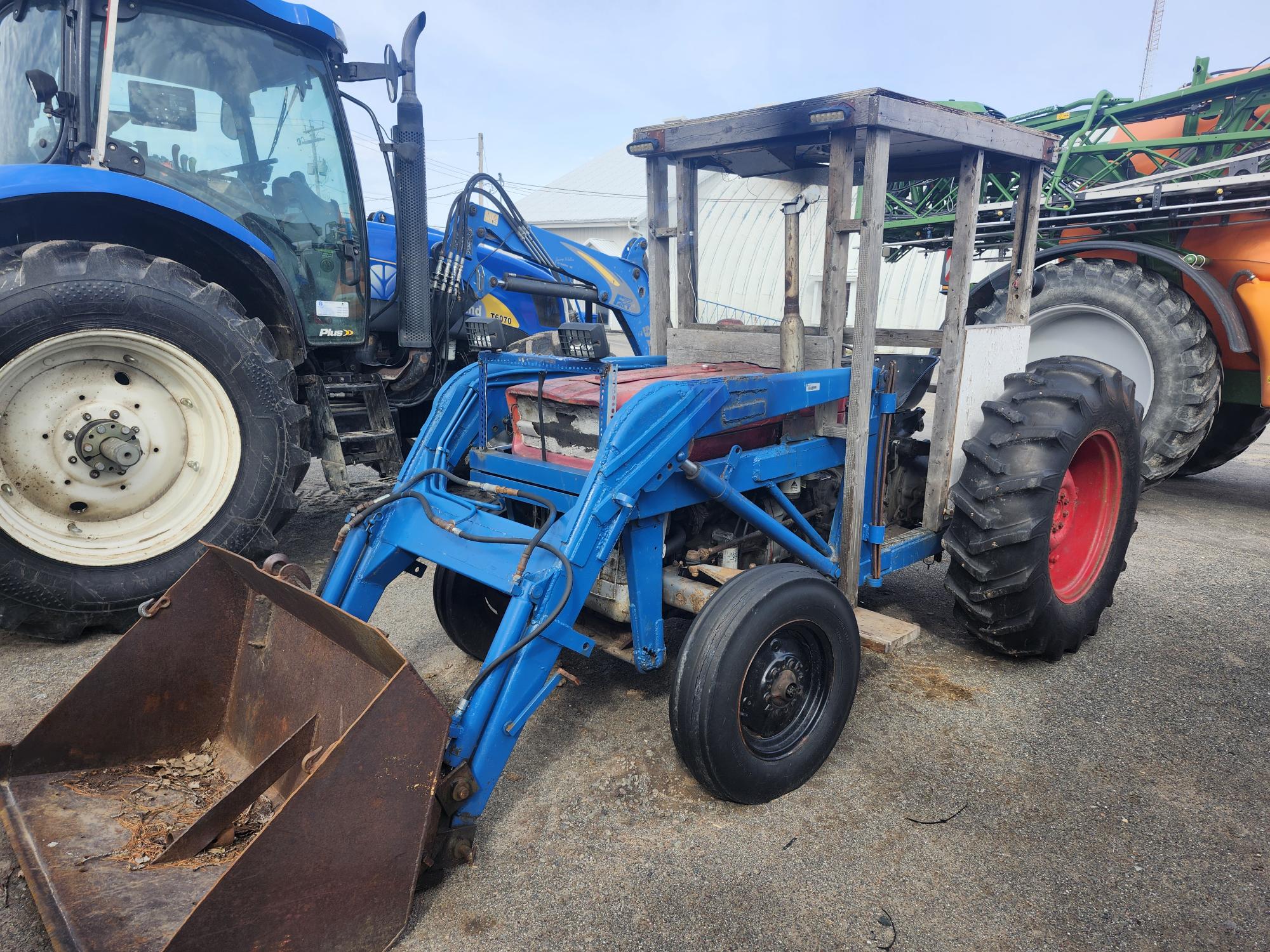 Un tracteur à chargeur frontal bleu et rouge avec une cabine à cadre métallique est garé sur le trottoir à côté d'un tracteur bleu plus gros et d'une machine agricole verte en arrière-plan.