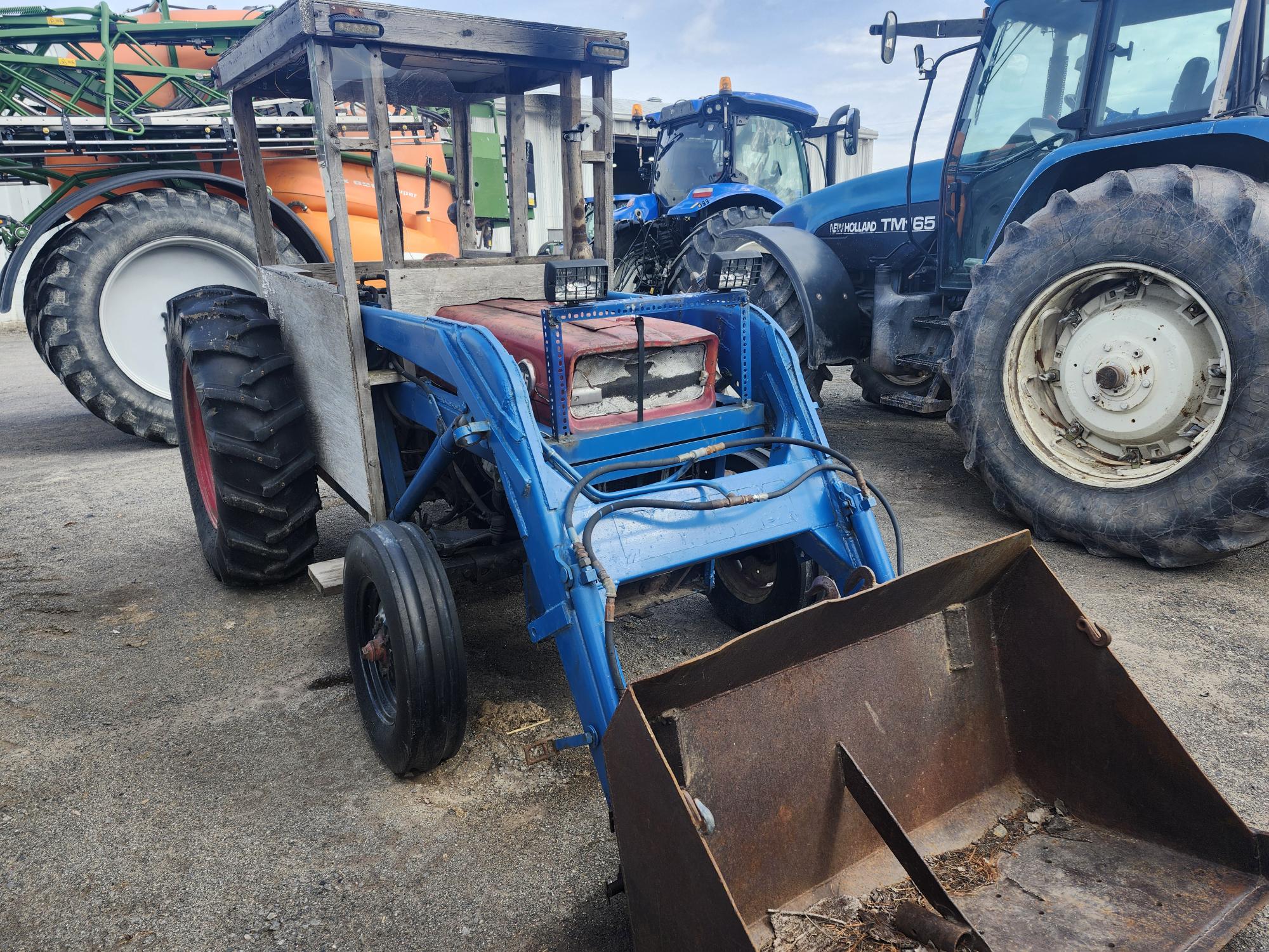 Un tracteur bleu équipé d'un chargeur frontal est garé sur une surface en béton parmi d'autres gros véhicules et équipements agricoles. Le tracteur paraît vieux et patiné.