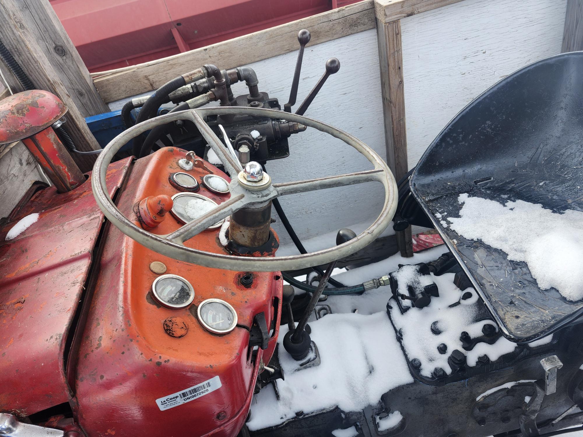 Un vieux tracteur avec un tableau de bord et un volant rouges et patinés. On y voit divers instruments. Le siège est recouvert de neige. Le tracteur est entouré de planches de bois et de pièces métalliques.