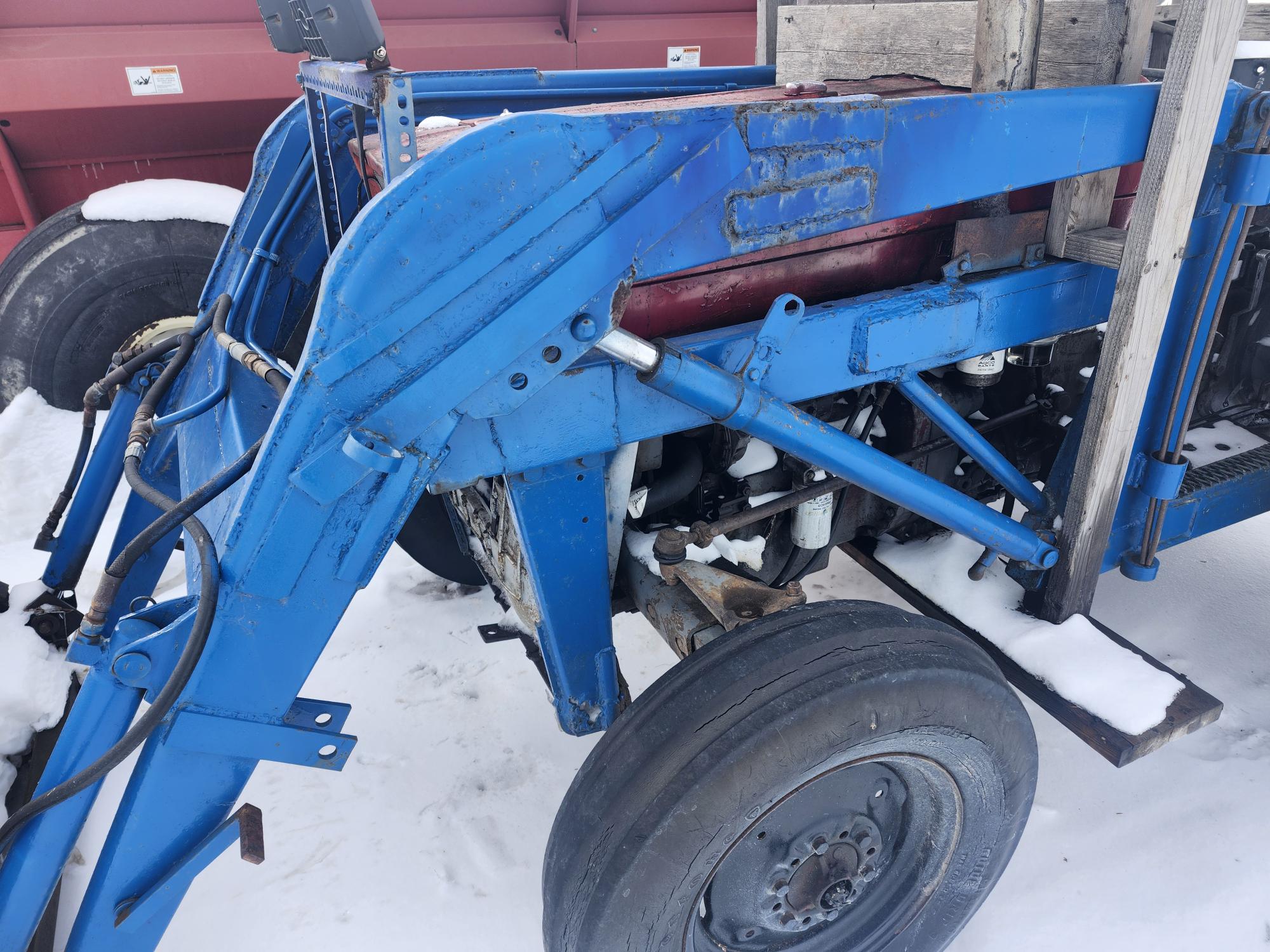 Un tracteur-chargeur bleu et usé dans une zone enneigée, avec des composants mécaniques visibles et une roue. Il est garé près d'un véhicule ou d'une remorque rouge.