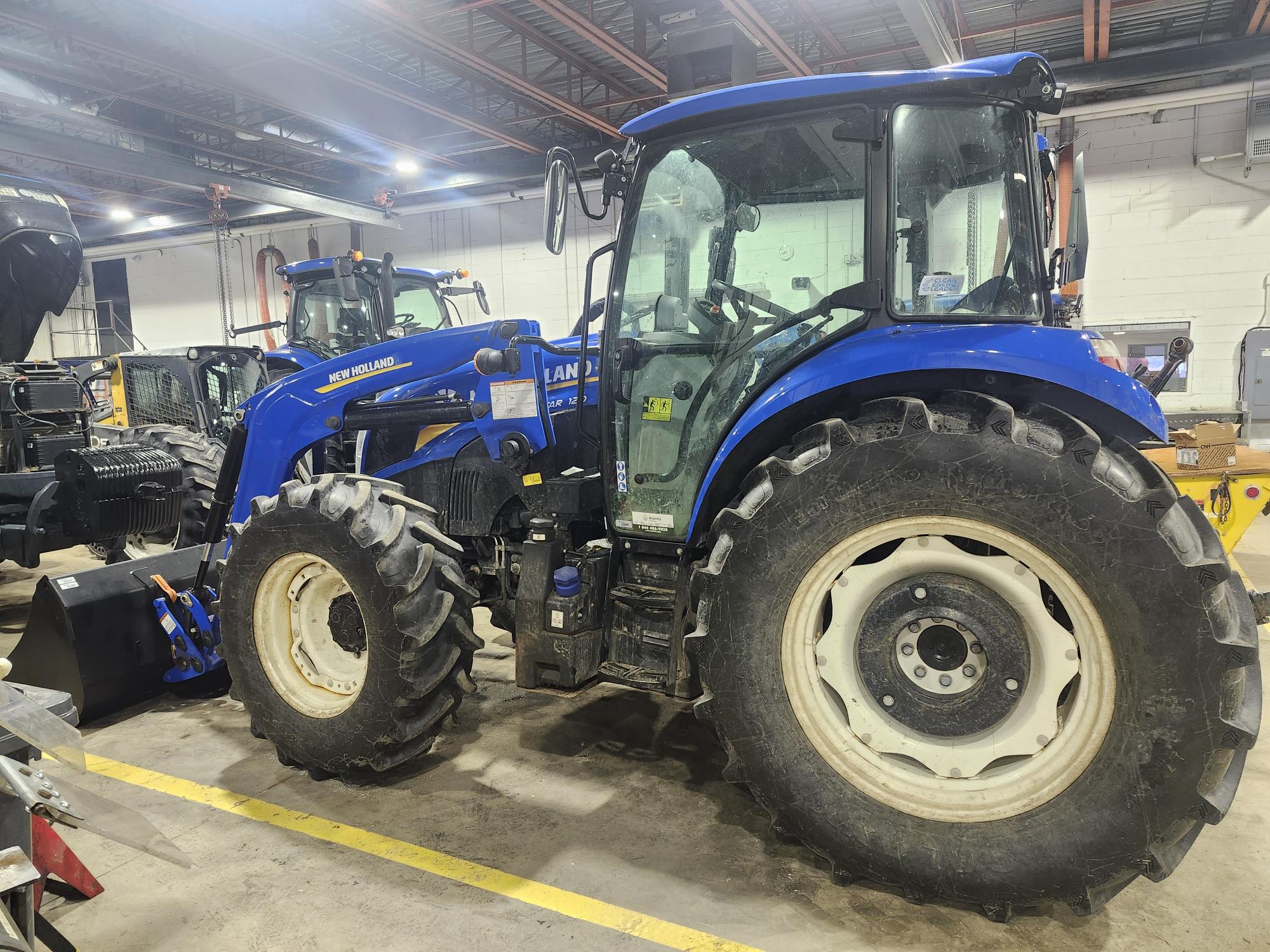 Un tracteur New Holland bleu est garé à l'intérieur sur un sol en béton. Il est équipé de gros pneus et d'un chargeur frontal. Le décor, rappelant celui d'un entrepôt, présente des étagères et des équipements industriels en arrière-plan.