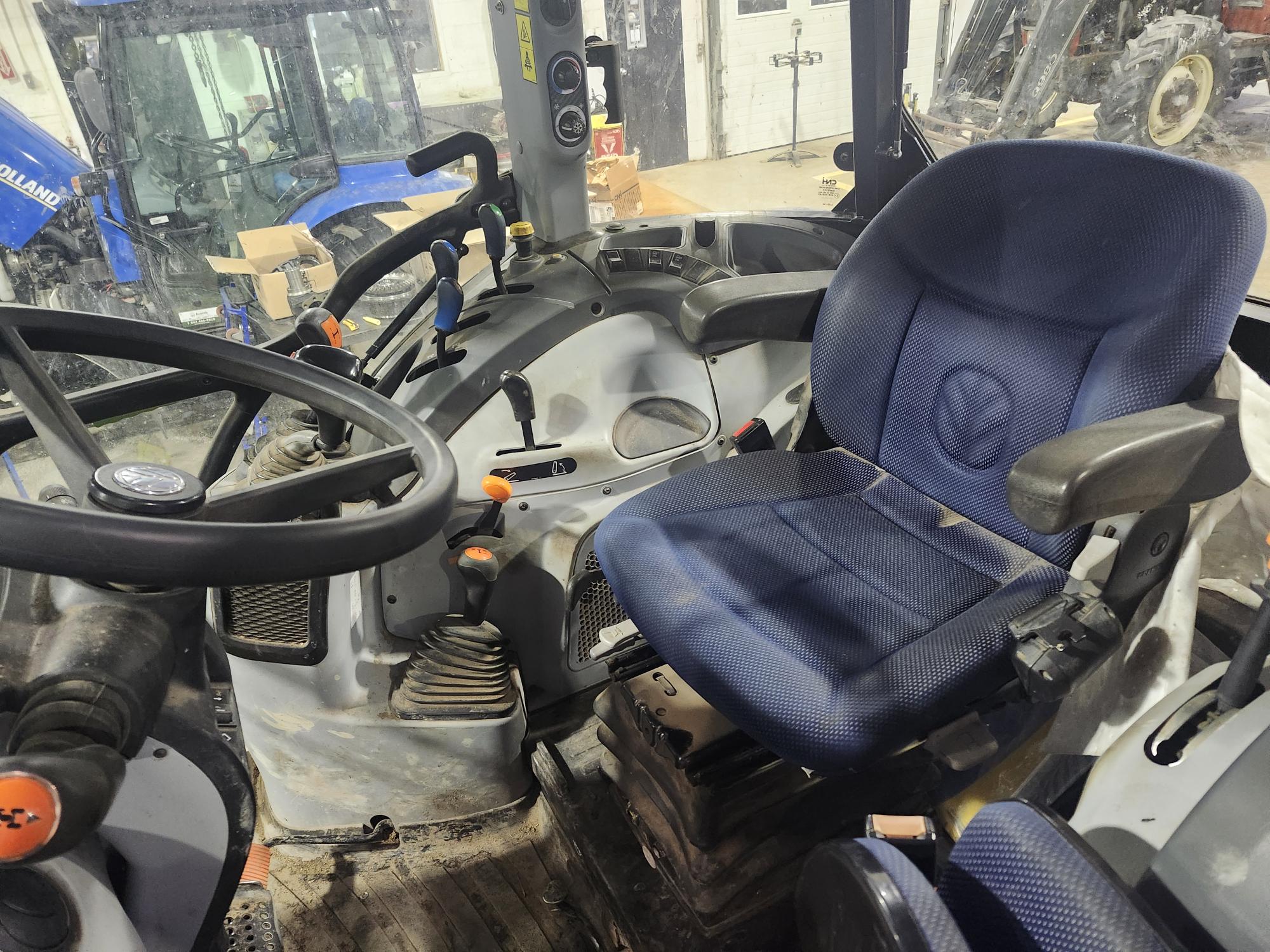 Intérieur d'une cabine de tracteur montrant un siège bleu rembourré, un volant, divers boutons et leviers de commande, ainsi qu'un sol légèrement sale. La cabine est bien éclairée et une partie d'un autre tracteur est visible par la fenêtre.