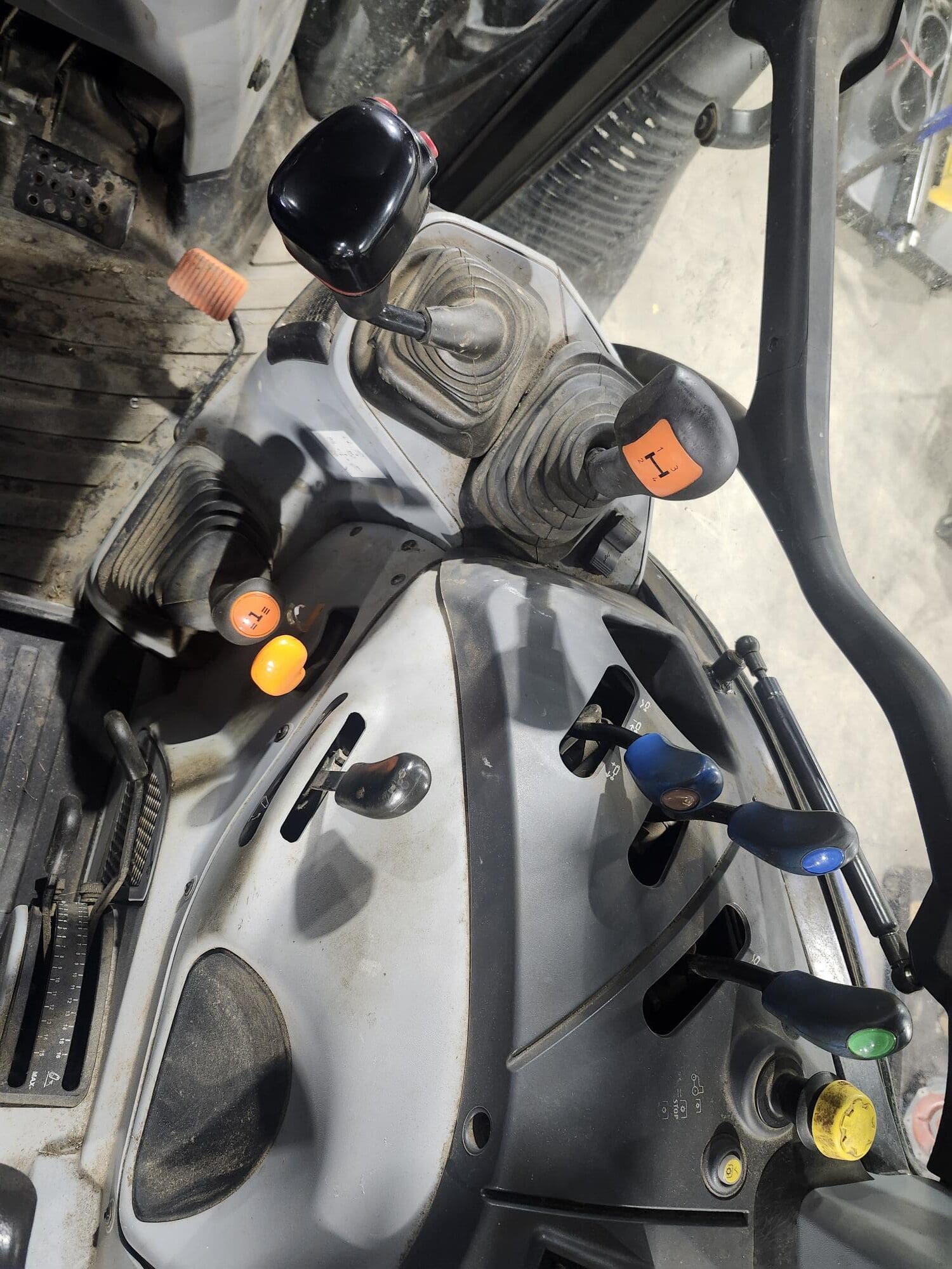 Intérieur d'un véhicule présentant diverses commandes, dont des leviers à boutons colorés, des pédales et un volant. Le sol est légèrement sale et la disposition suggère un usage industriel ou agricole.