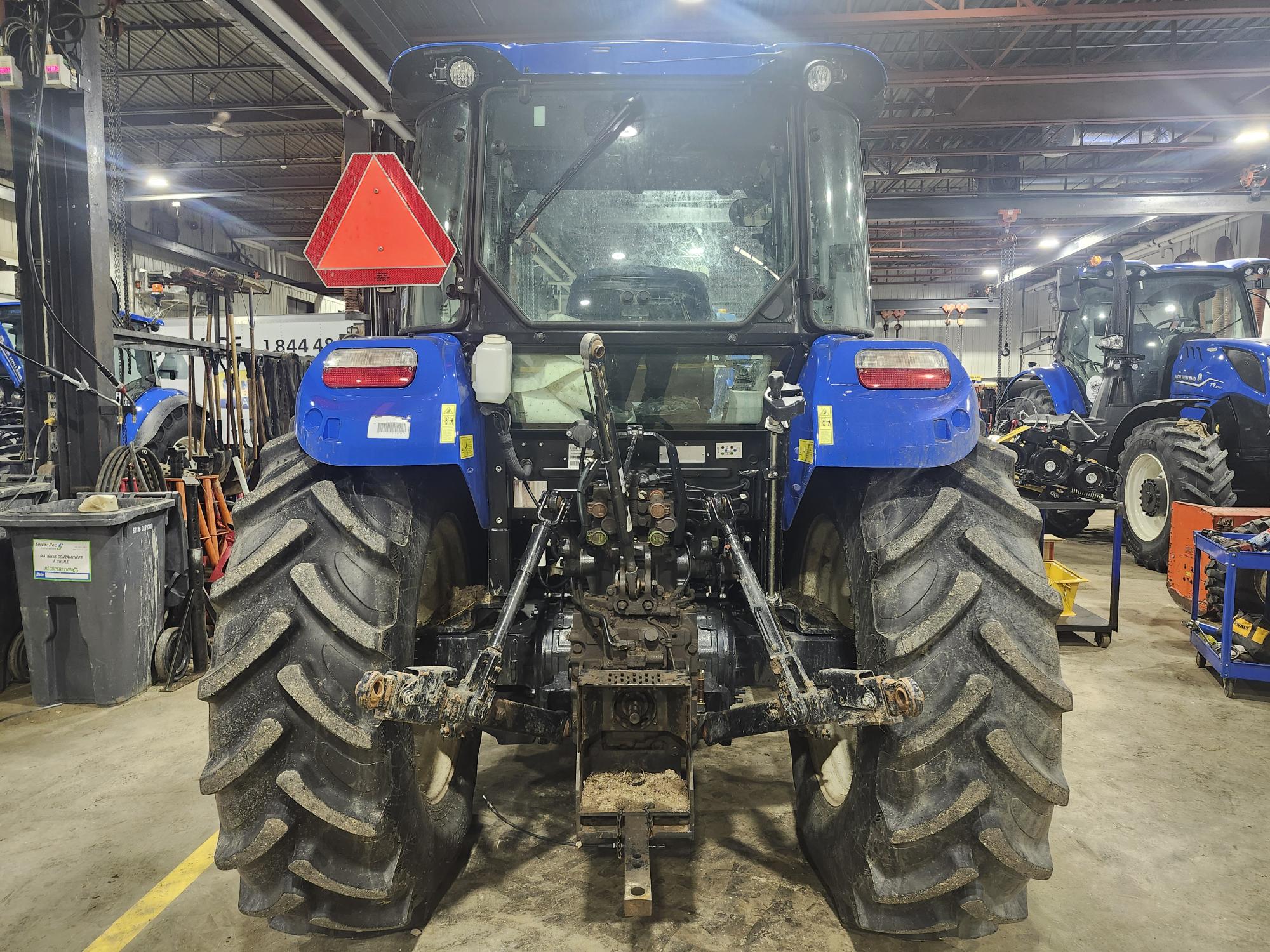 Un tracteur bleu est garé dans un atelier industriel. Il est équipé de grandes roues arrière et d'un panneau d'avertissement triangulaire orange fixé à l'arrière. L'atelier présente divers outils et d'autres tracteurs en arrière-plan.