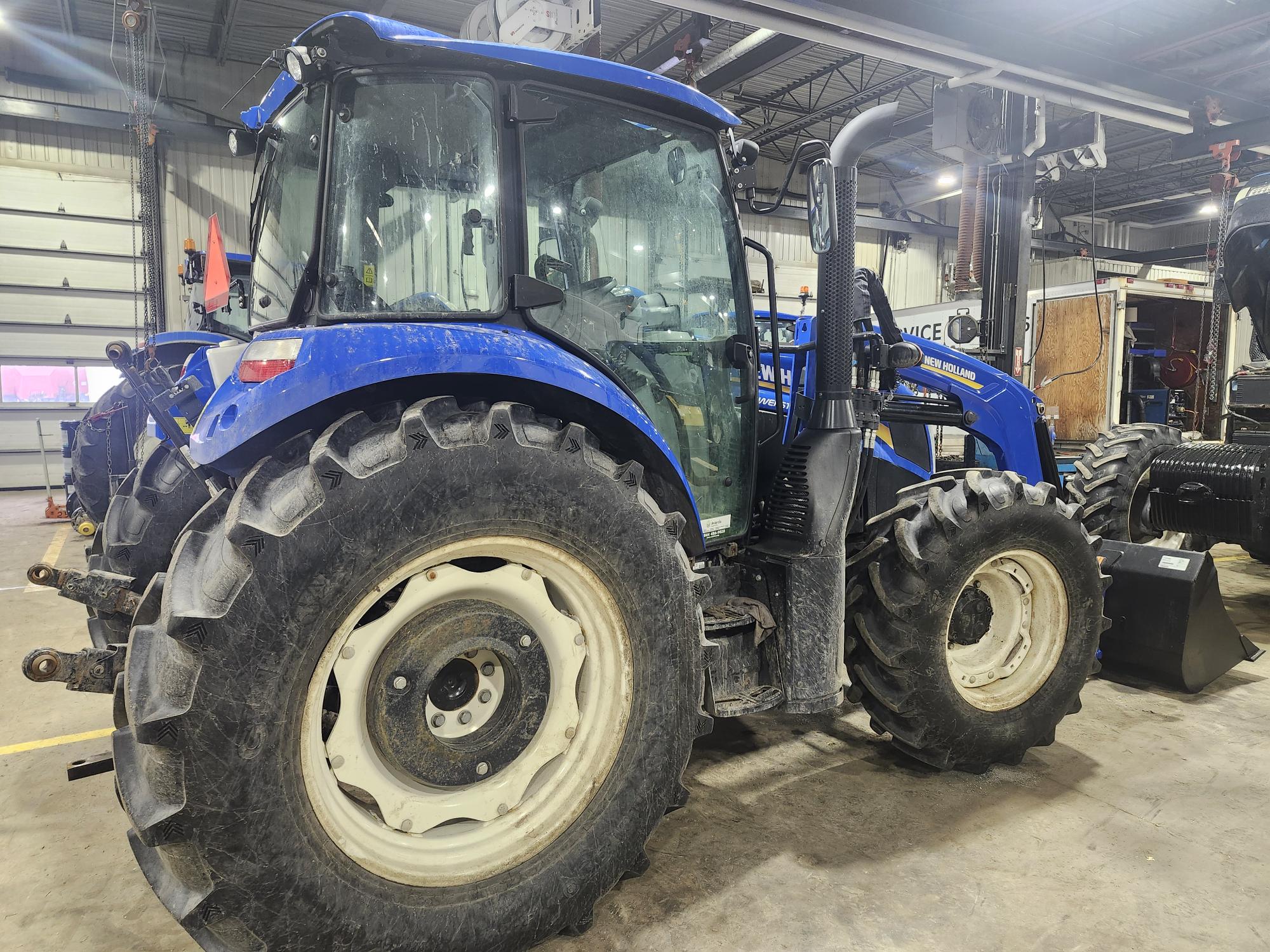 Tracteur bleu avec de gros pneus noirs, garé à l'intérieur d'un garage ou d'un atelier. Le véhicule possède une cabine vitrée et un pot d'échappement visible. L'arrière-plan révèle des équipements de garage et une autre pièce du véhicule.