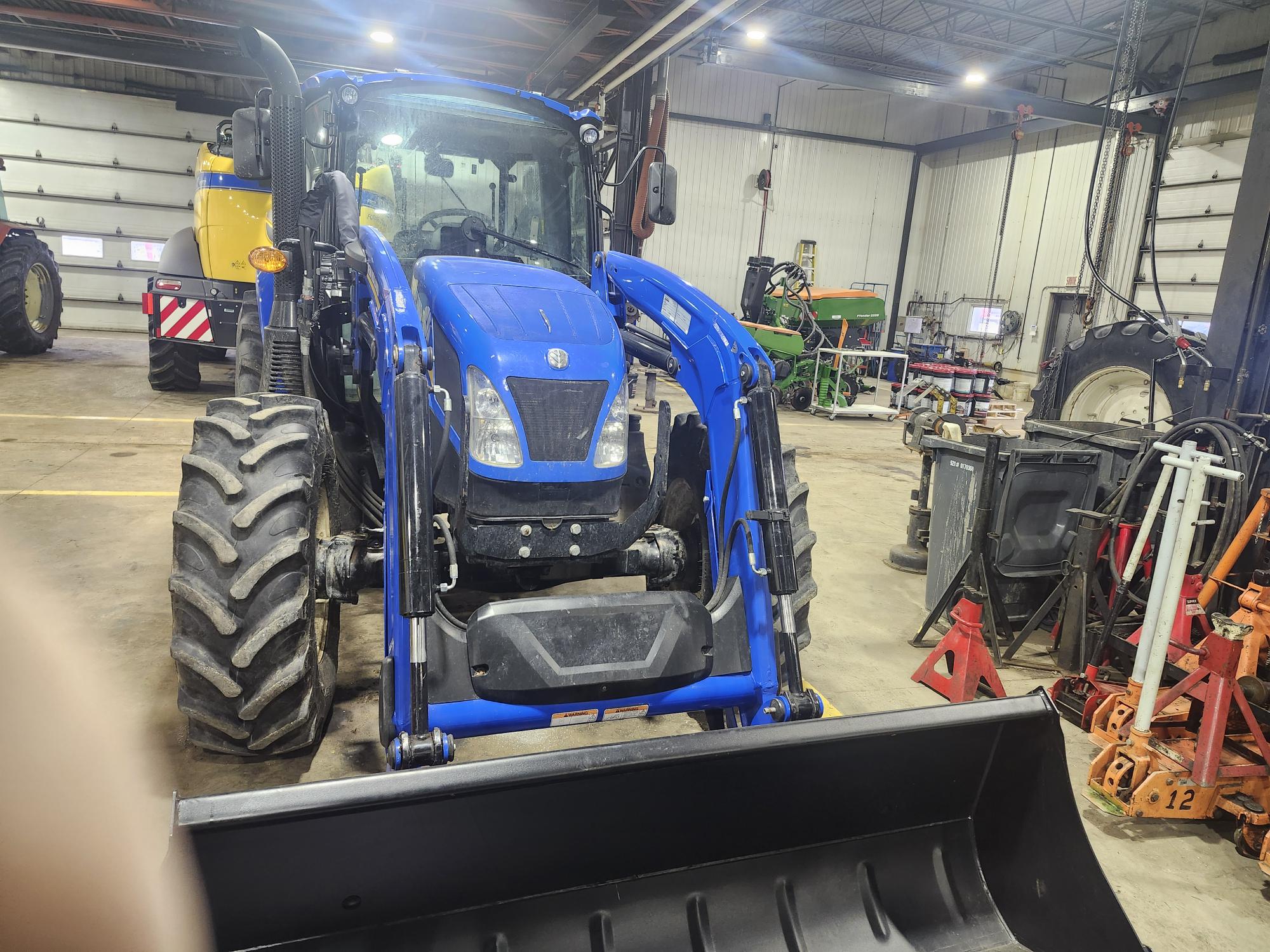 Tracteur bleu avec chargeur frontal garé dans un garage spacieux. L'environnement comprend divers équipements et machines agricoles, avec un sol en béton et un haut plafond agrémenté de lumières.