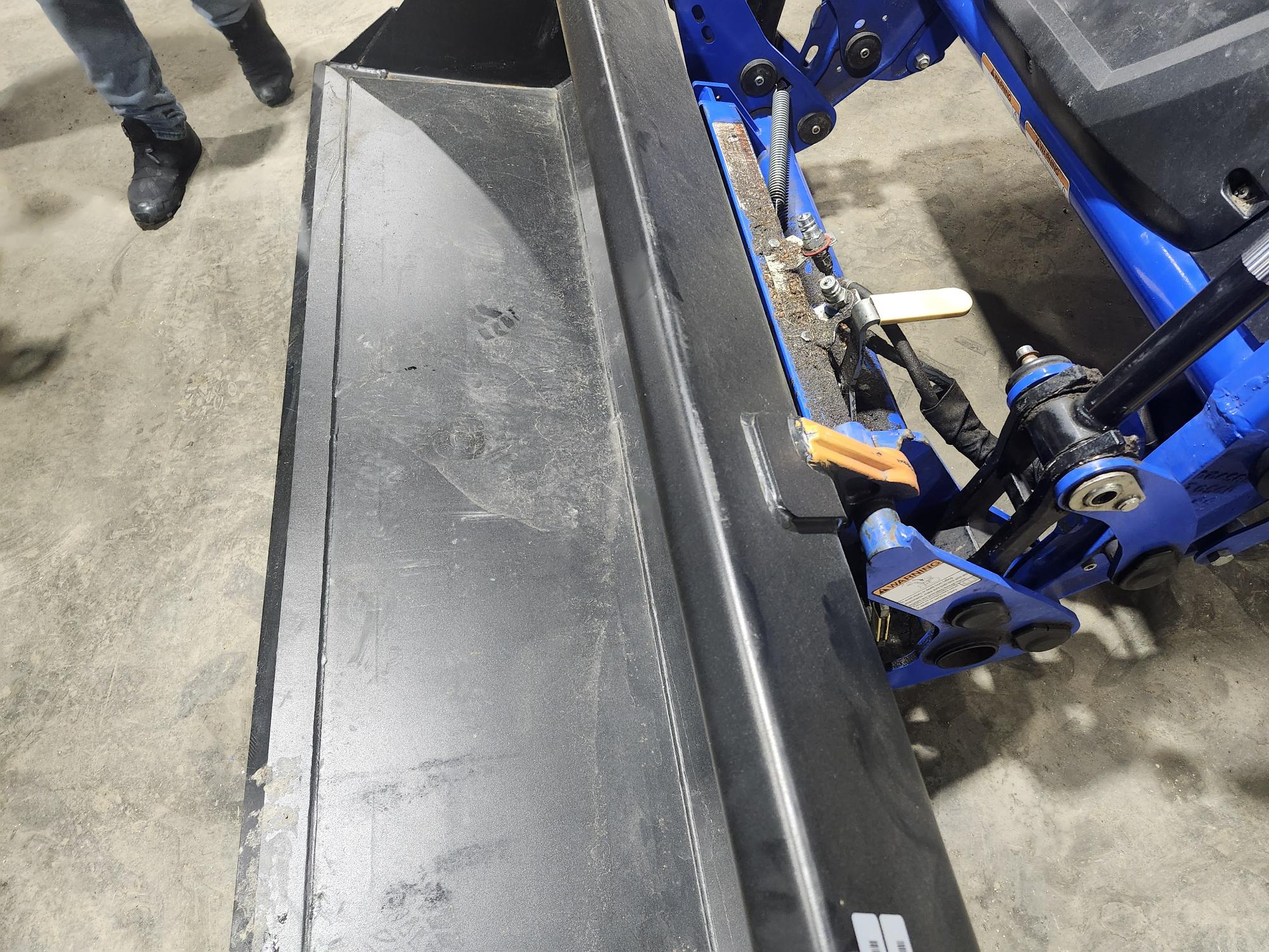 Gros plan d'un grand chasse-neige industriel attaché à une machine bleue, vu de côté. Le sol est en béton, et on aperçoit les jambes et les pieds d'une personne sur le côté gauche.