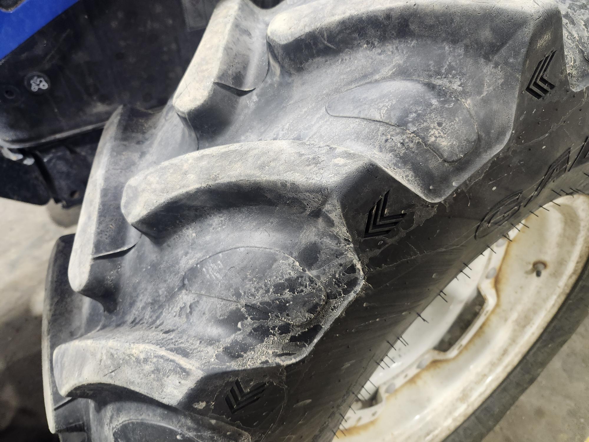 Gros plan sur un gros pneu de tracteur robuste aux sculptures profondes. Le pneu est fixé à une jante blanche, partiellement visible. La surface du pneu présente des traces de poussière et d'usure, témoignant d'une utilisation prolongée. L'arrière-plan est un sol en béton gris.