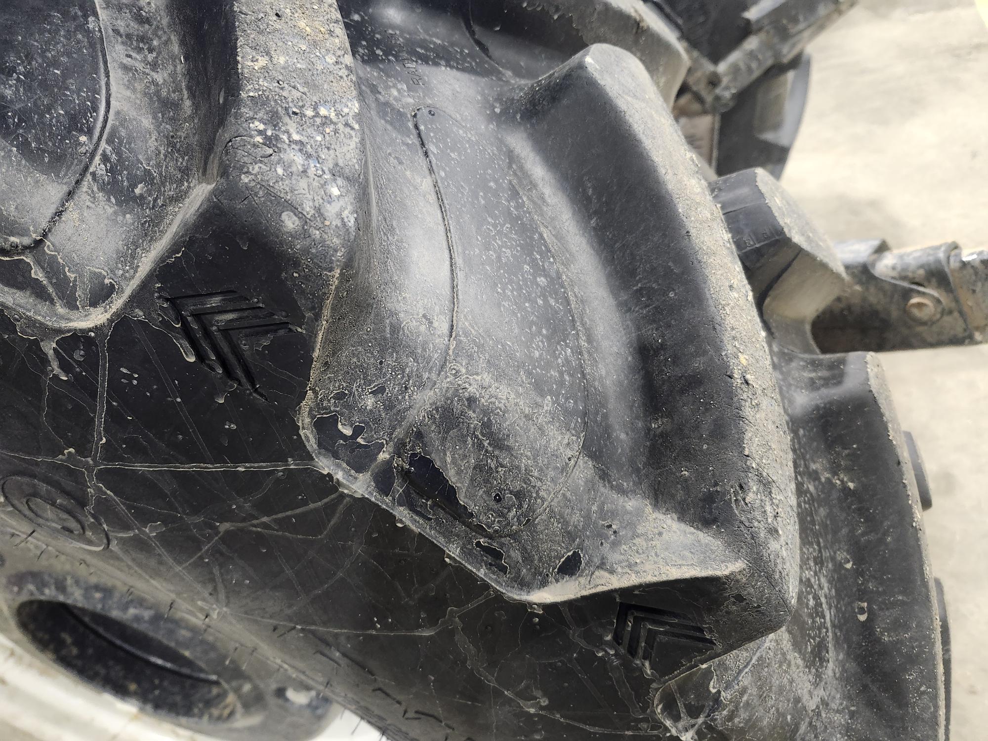 Gros plan d'un pneu de tracteur usé présentant d'importantes fissures et dommages sur la surface en caoutchouc, ainsi que des traces de saleté visibles. La sculpture du pneu est prononcée et certains détails structurels sont visibles.
