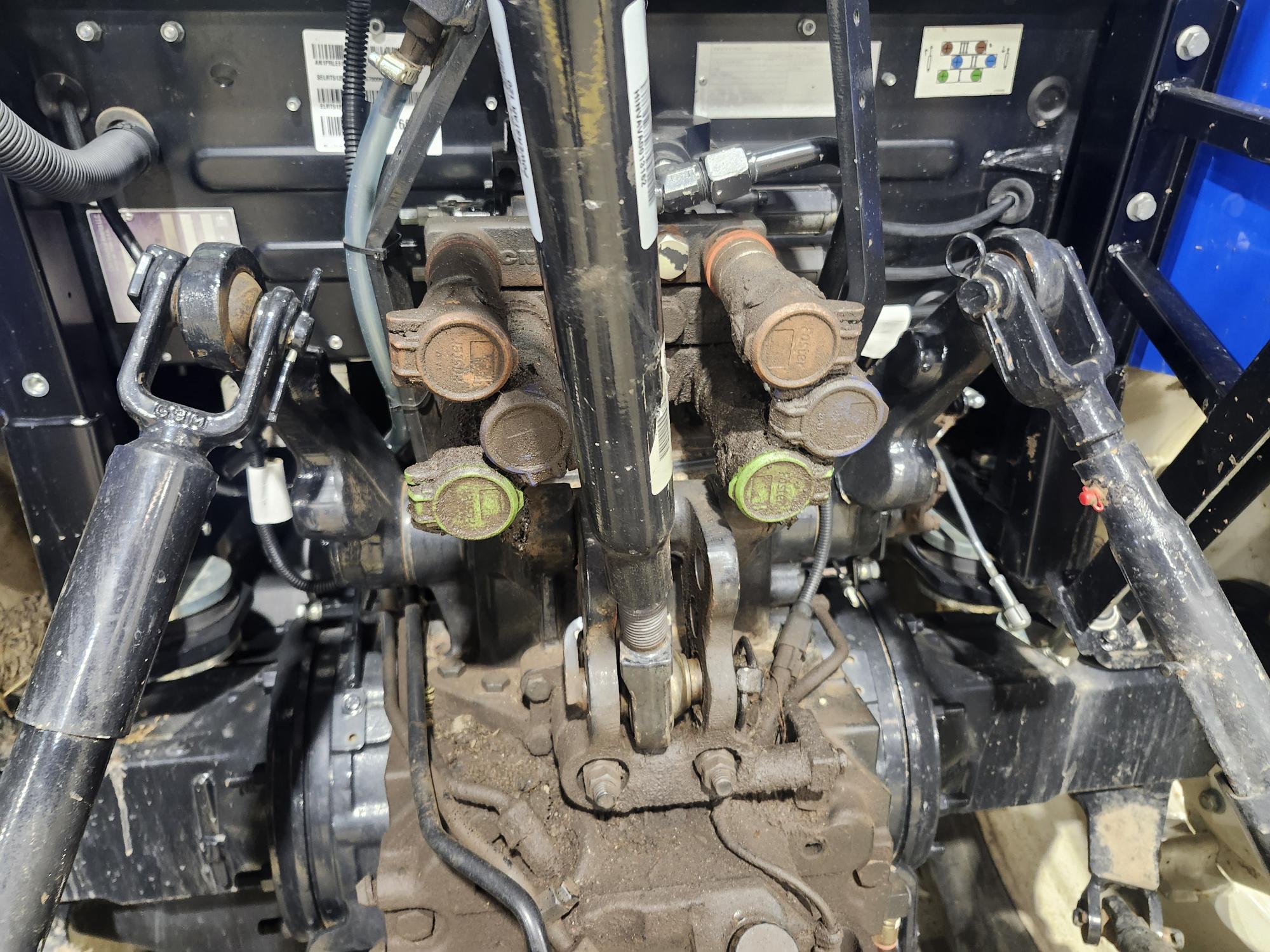 Gros plan du système hydraulique d'un tracteur, avec ses multiples raccords et flexibles hydrauliques à code couleur. La machine est recouverte de terre et diverses pièces métalliques sont visibles, soulignant la complexité des composants.