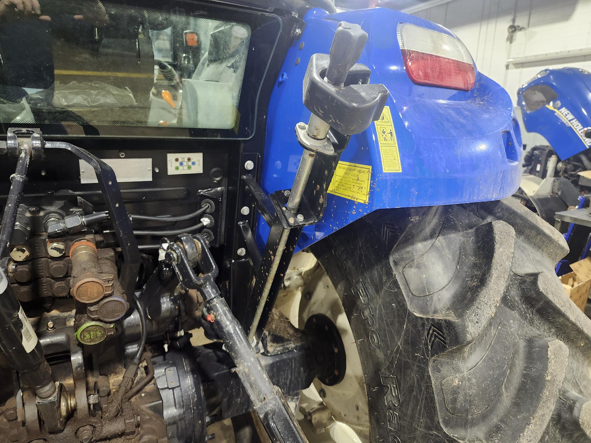 Un tracteur bleu dans un atelier, montrant sa roue arrière, son système hydraulique et son levier de commande. Le tracteur est posé sur un sol en béton, avec un autre tracteur visible en arrière-plan.